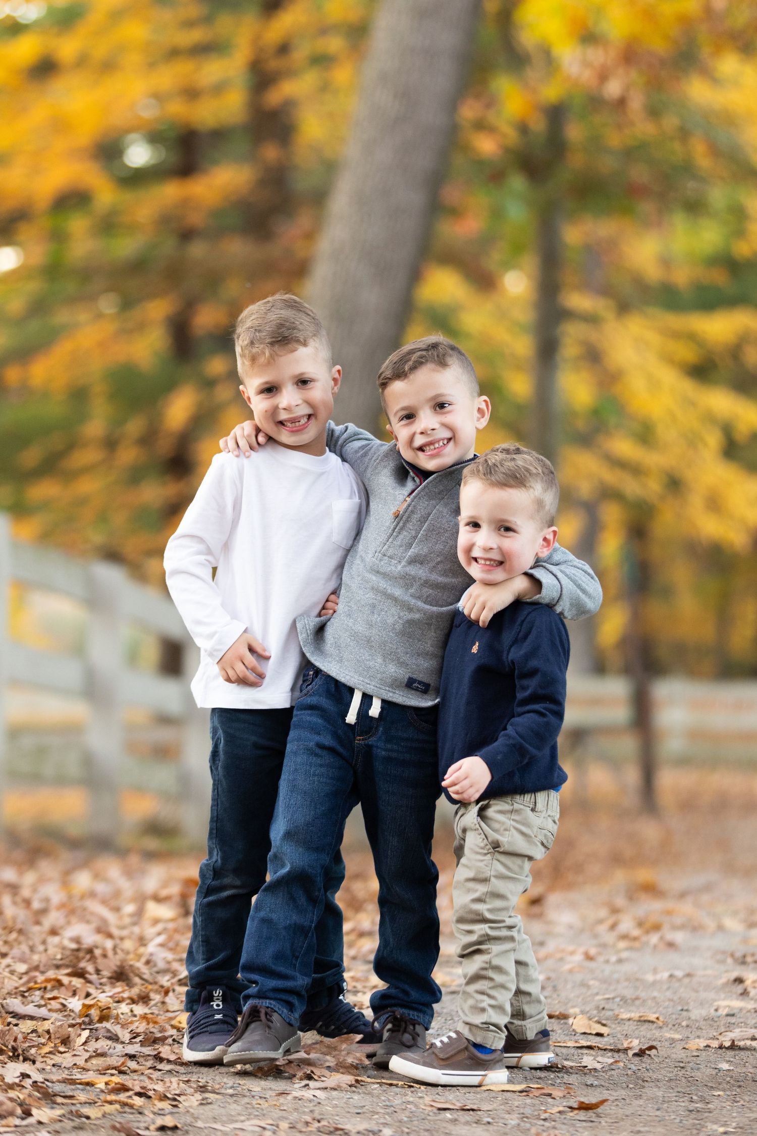 Three young brothers hugging and smiling together on a path surrounded by colorful autumn foliage.