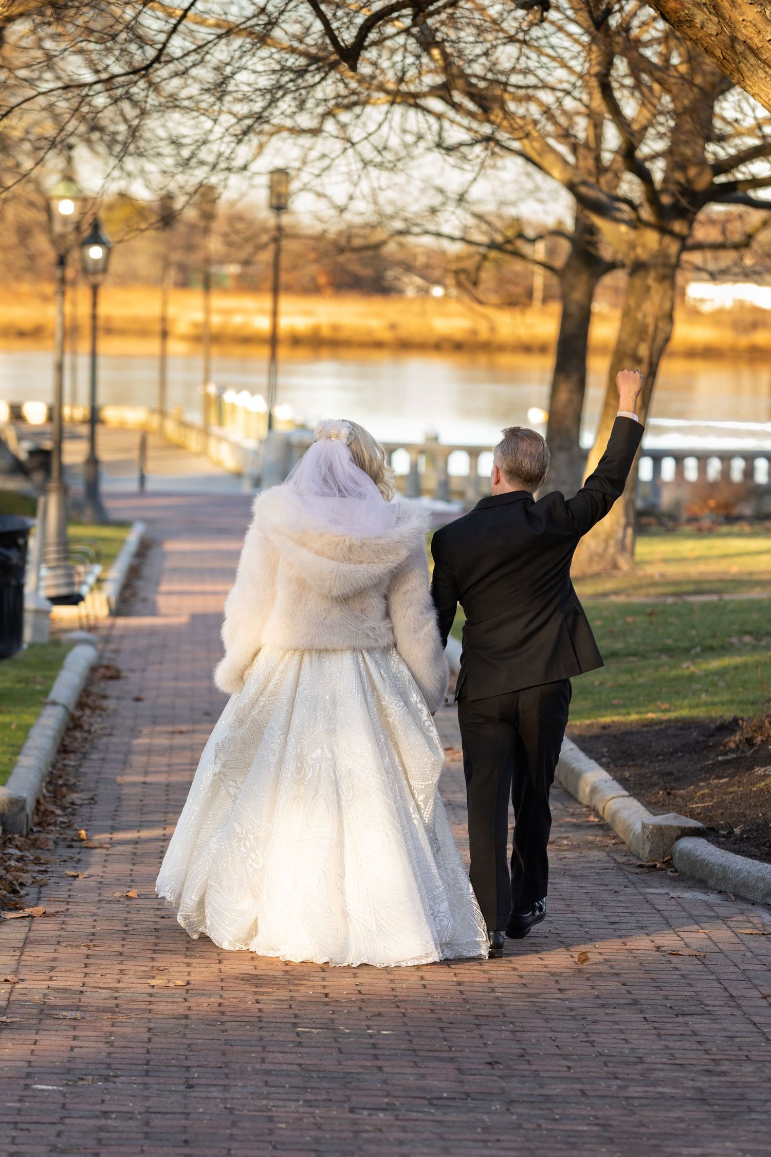 Bride in white gown and fur wrap walks with groom in black suit along brick path by river, groom raising hand.