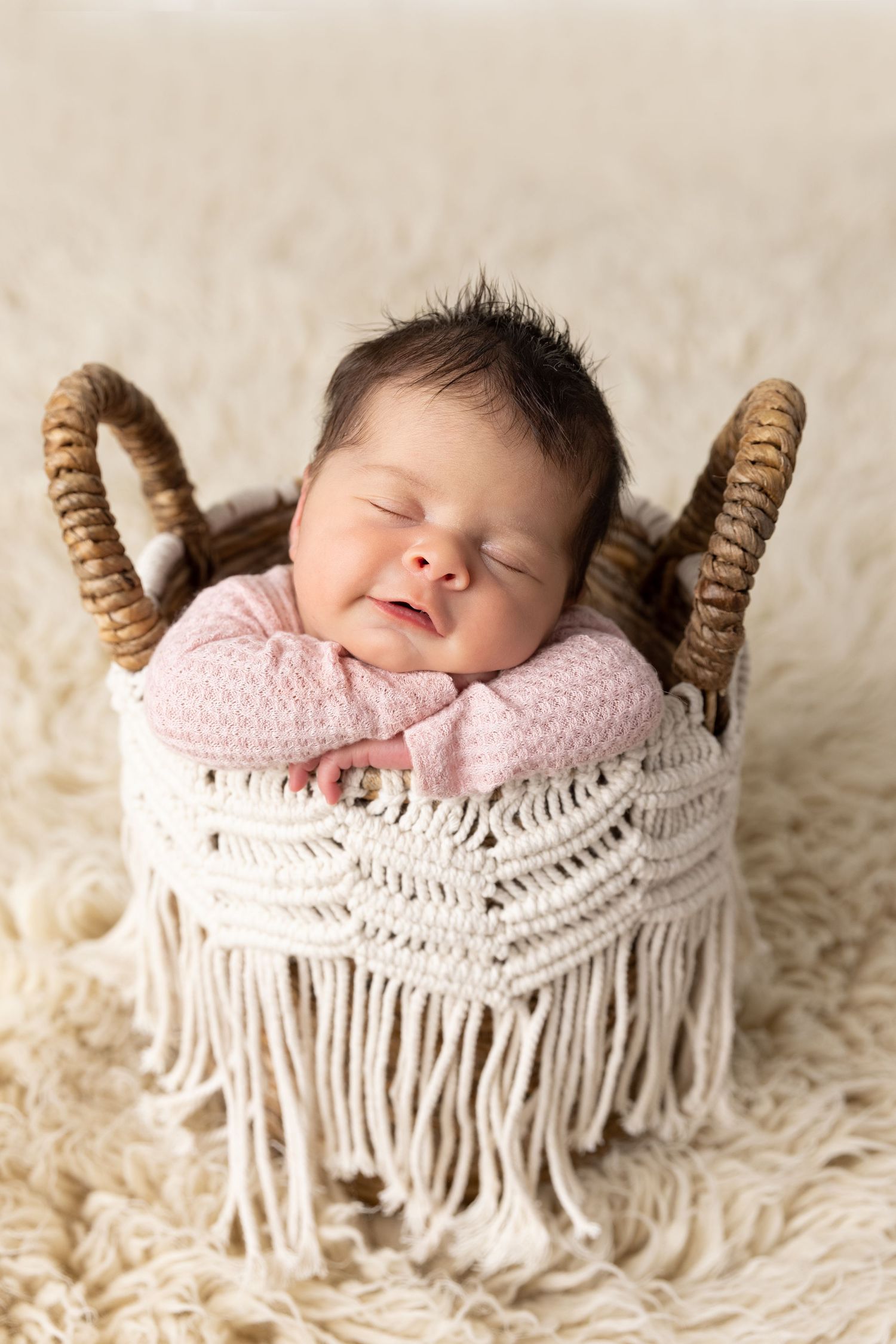 Smiling newborn baby sleeping in a white macrame basket with fringe, wrapped in a pink swaddle on a cream fluffy rug.