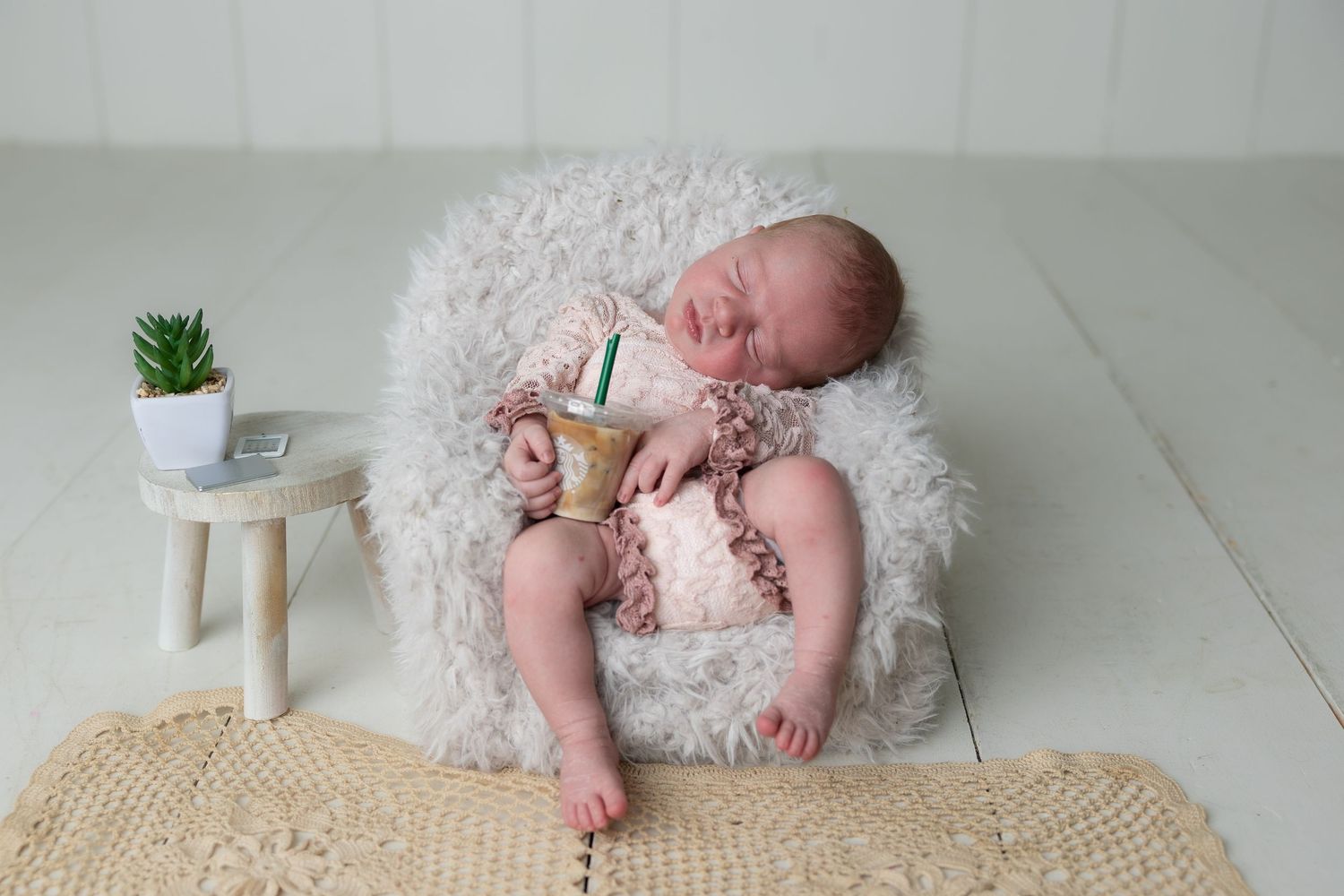 Newborn baby sleeping on a fluffy white chair holding a mini Starbucks cup, with a small plant and books nearby.