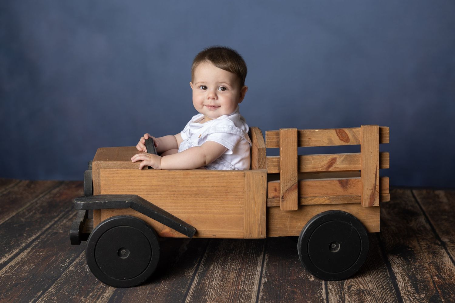 Smiling baby sitting in a wooden toy truck on rustic hardwood floors against a blue backdrop.