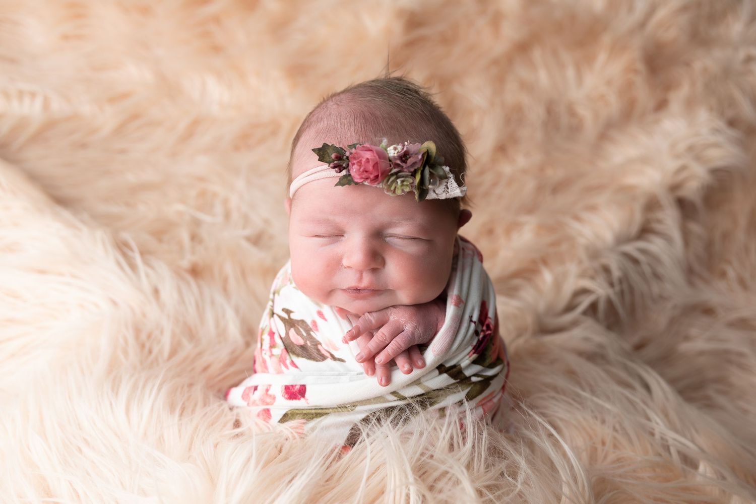 Sleeping newborn baby swaddled in floral wrap with pink flower headband on fluffy cream fur backdrop.