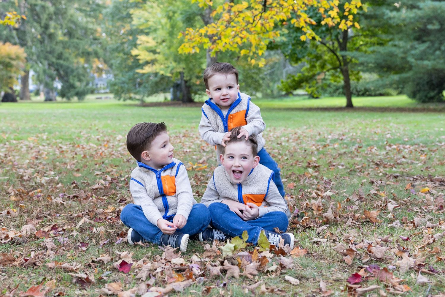 Three young siblings in matching outfits playing in autumn leaves at a park.