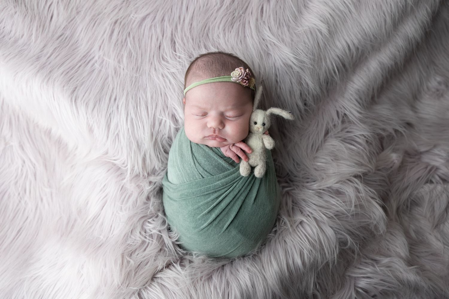 Newborn baby swaddled in green wrap with bunny toy, sleeping on fluffy white fur backdrop.
