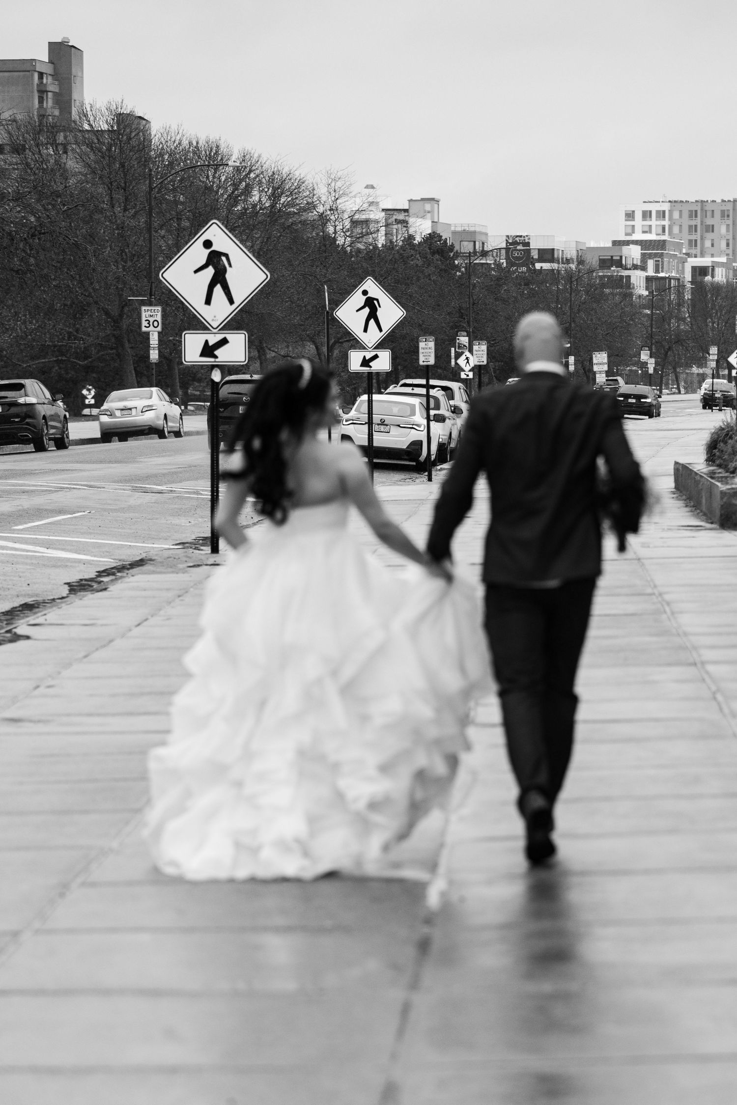 Black and white photo of a bride and groom walking hand in hand on a city sidewalk, bride in a ruffled gown.