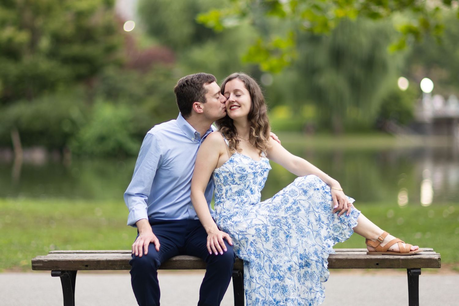 Couple sharing a romantic moment on a park bench near a pond, woman in blue floral dress, man in light blue shirt.