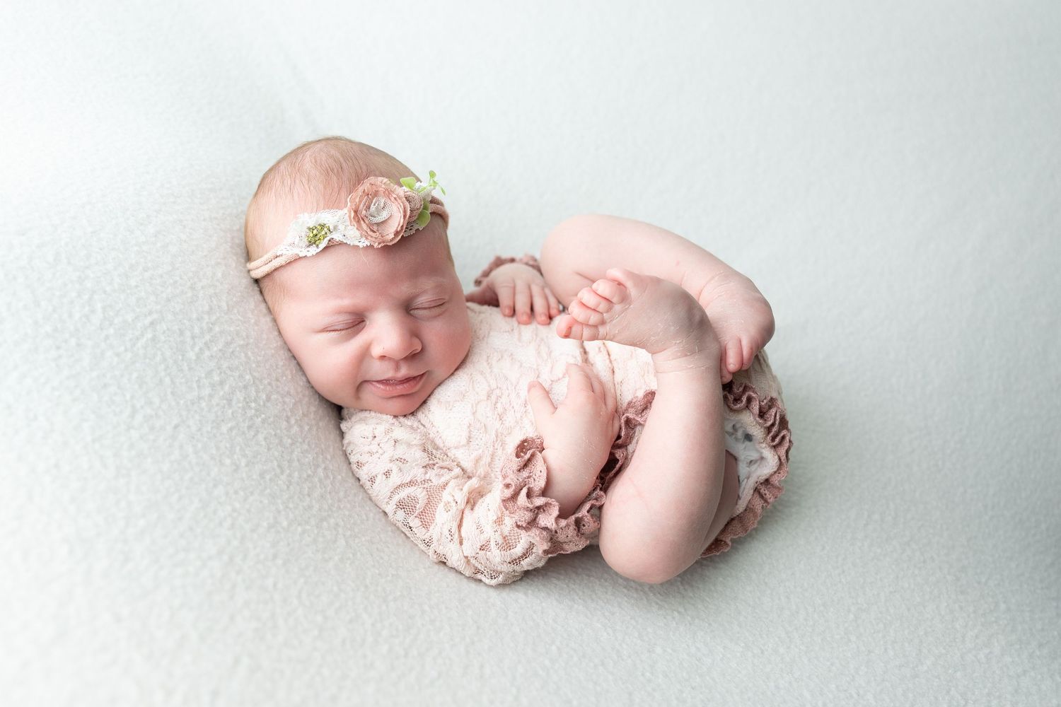 Sleeping newborn baby girl in pink lace romper and floral headband on mint green backdrop.