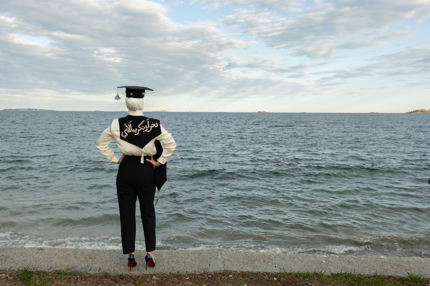 Graduate in cap and gown stands facing the ocean shore on a cloudy day.