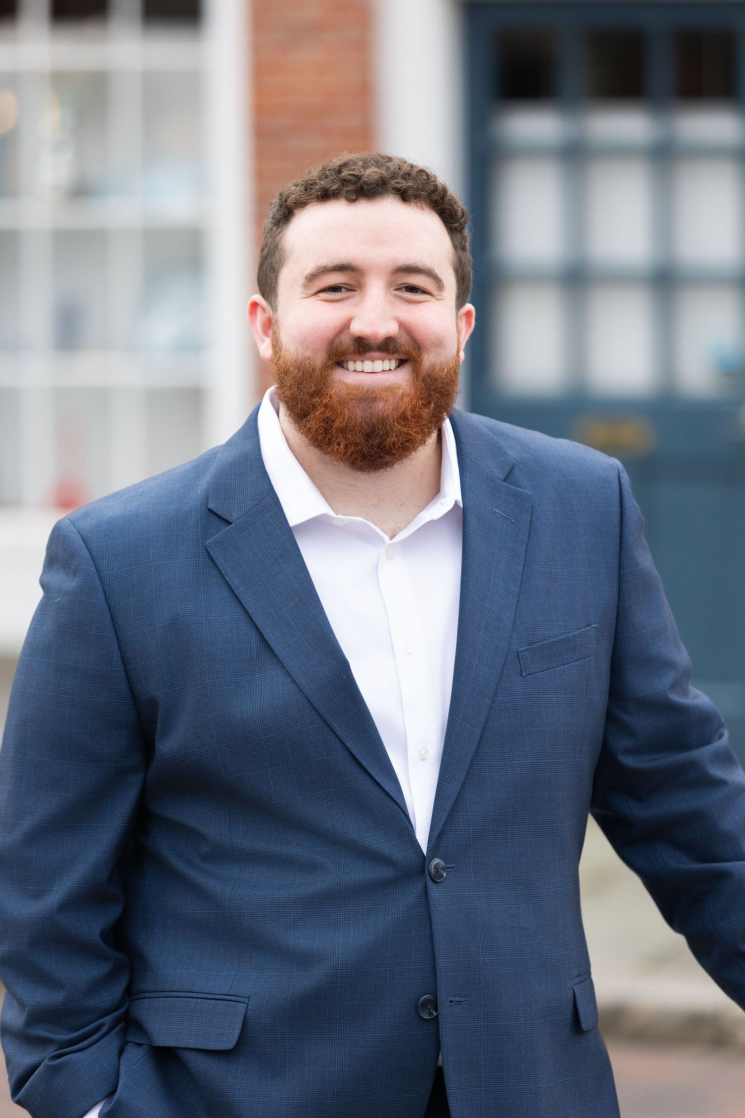 Smiling man with red beard wearing navy blue blazer and white shirt standing outdoors near brick building.