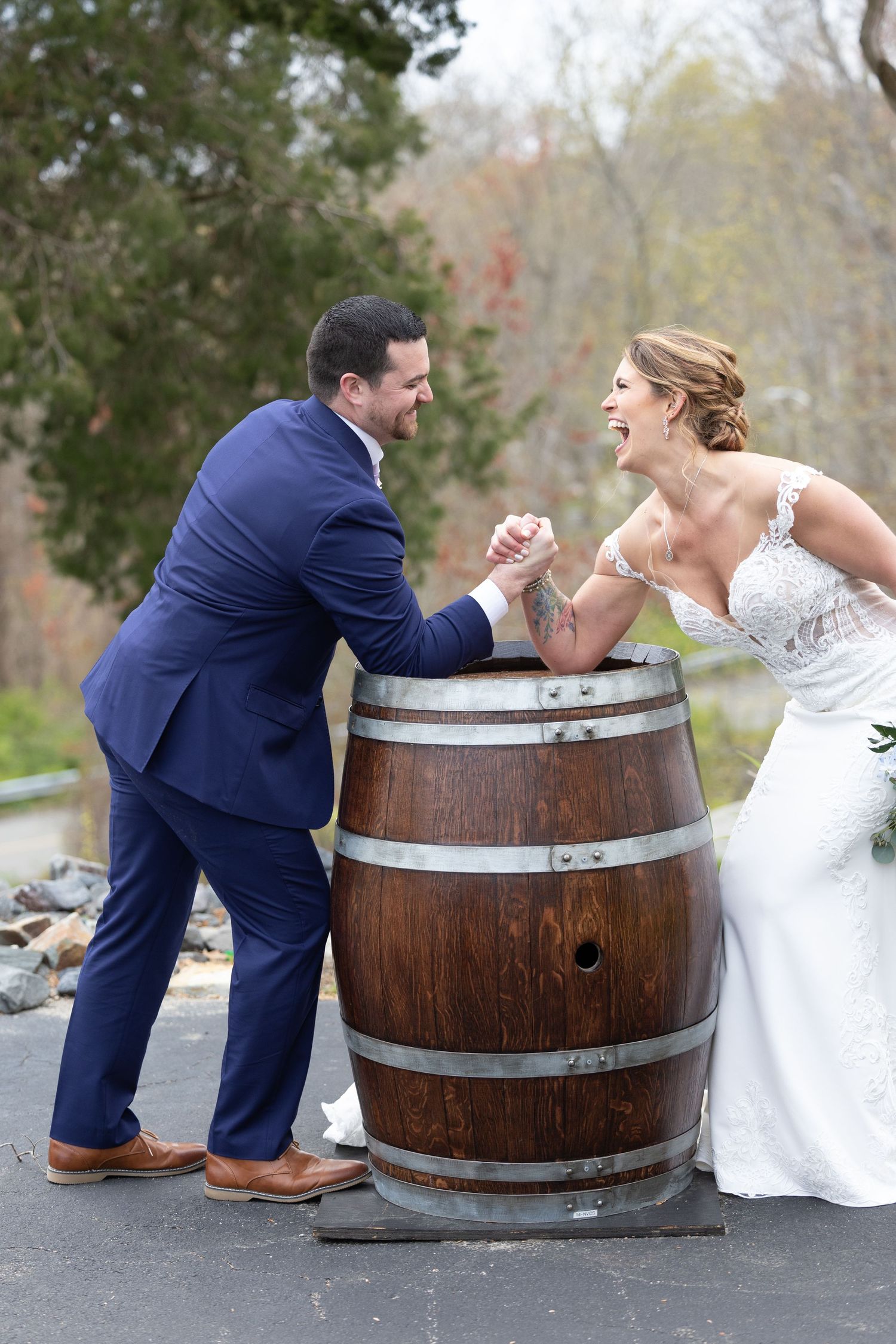 Bride and groom arm wrestling over a wooden wine barrel, laughing joyfully on their wedding day.