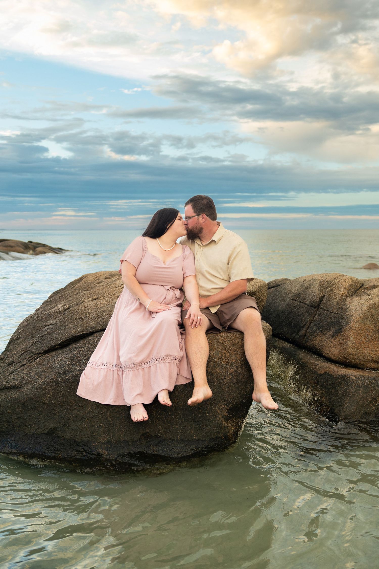 Couple sitting on coastal rocks kissing at sunset, woman in pink dress, man in khaki shirt, ocean backdrop.