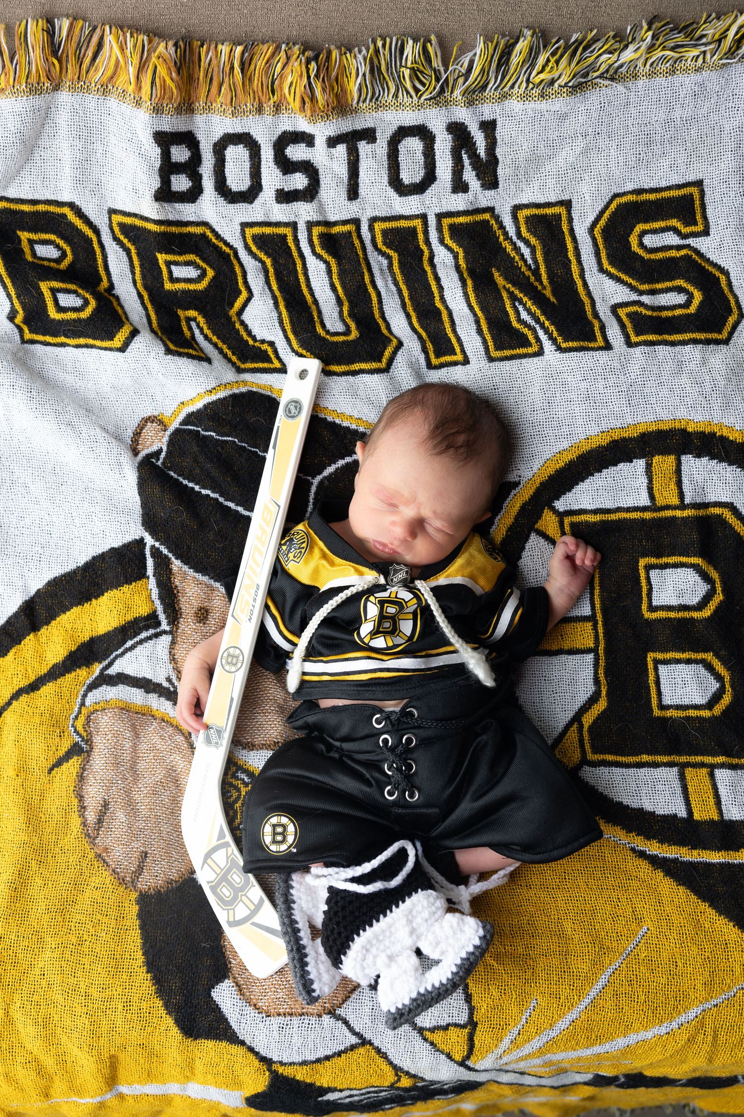 Newborn baby sleeping on a Boston Bruins blanket wearing a Bruins jersey outfit with a tiny hockey stick.