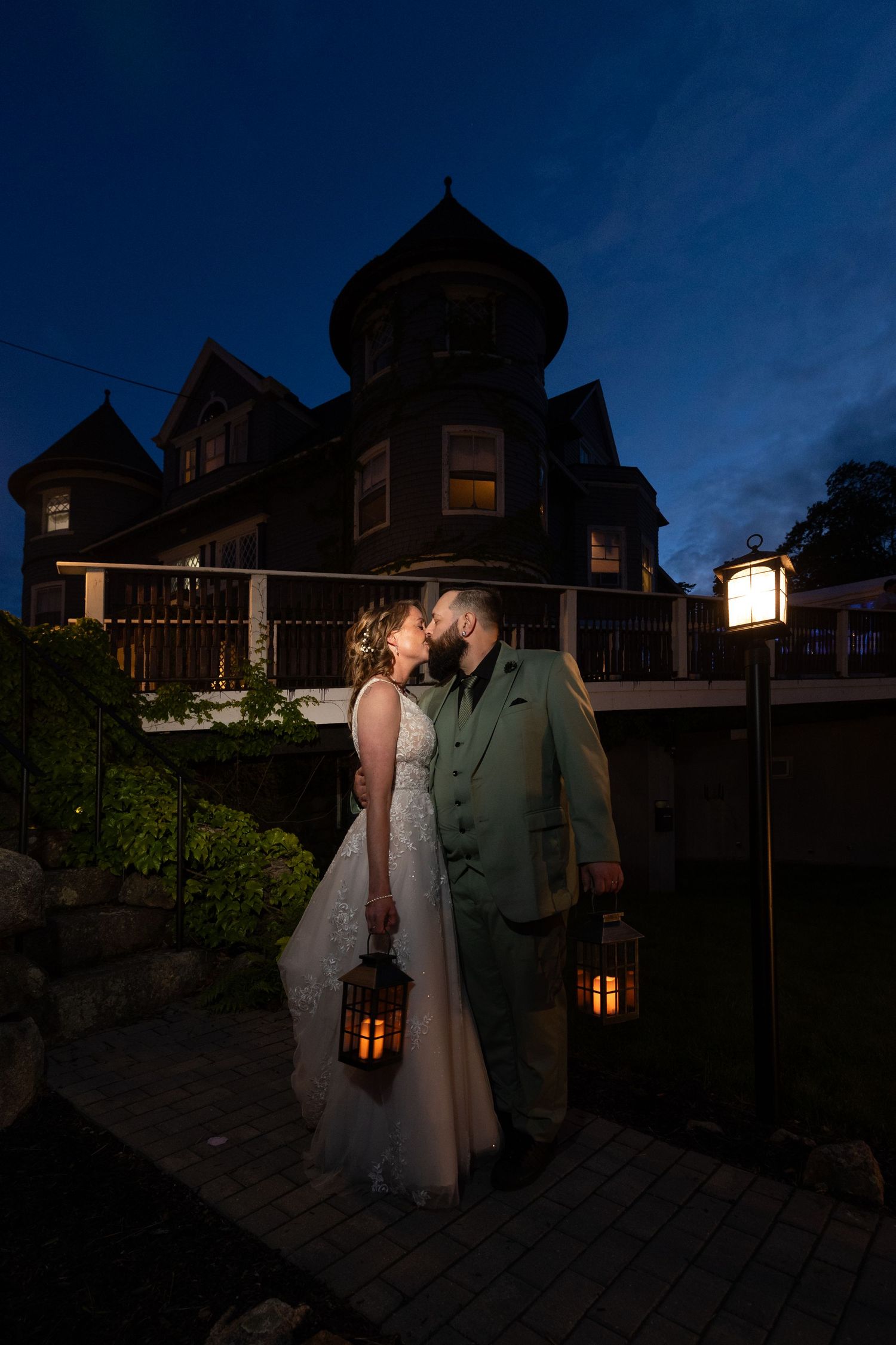 Bride and groom kissing at twilight holding lanterns before a Victorian mansion.
