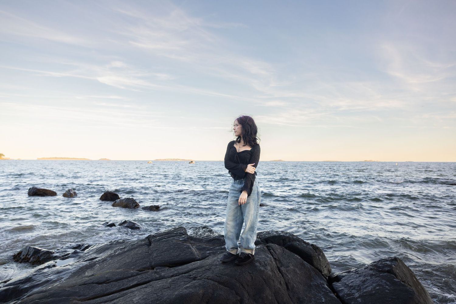 A young woman in jeans and black top stands on coastal rocks gazing at the ocean during golden hour.