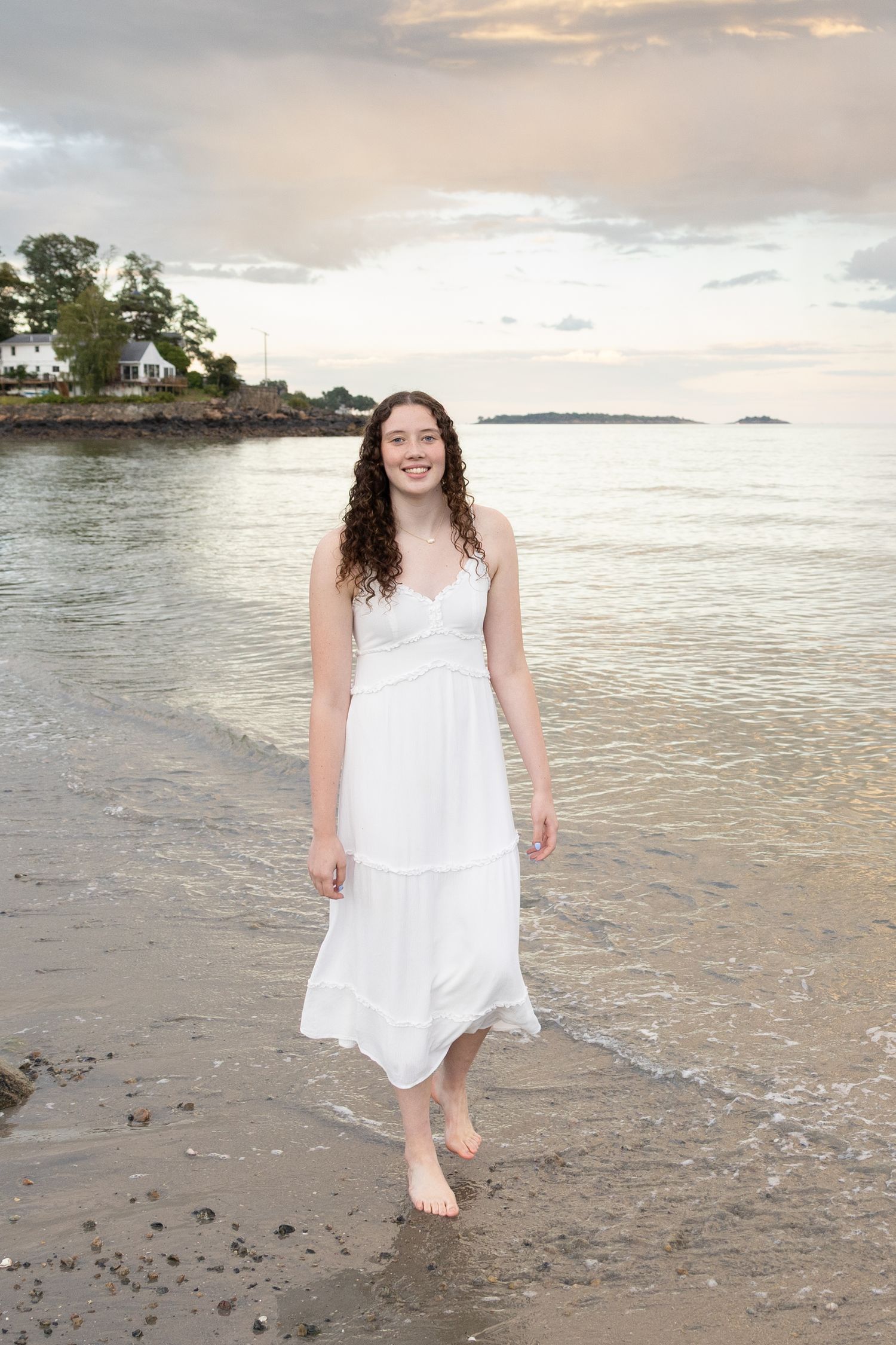 Young woman in white sundress smiling on sandy beach at sunset, waves lapping at her bare feet.