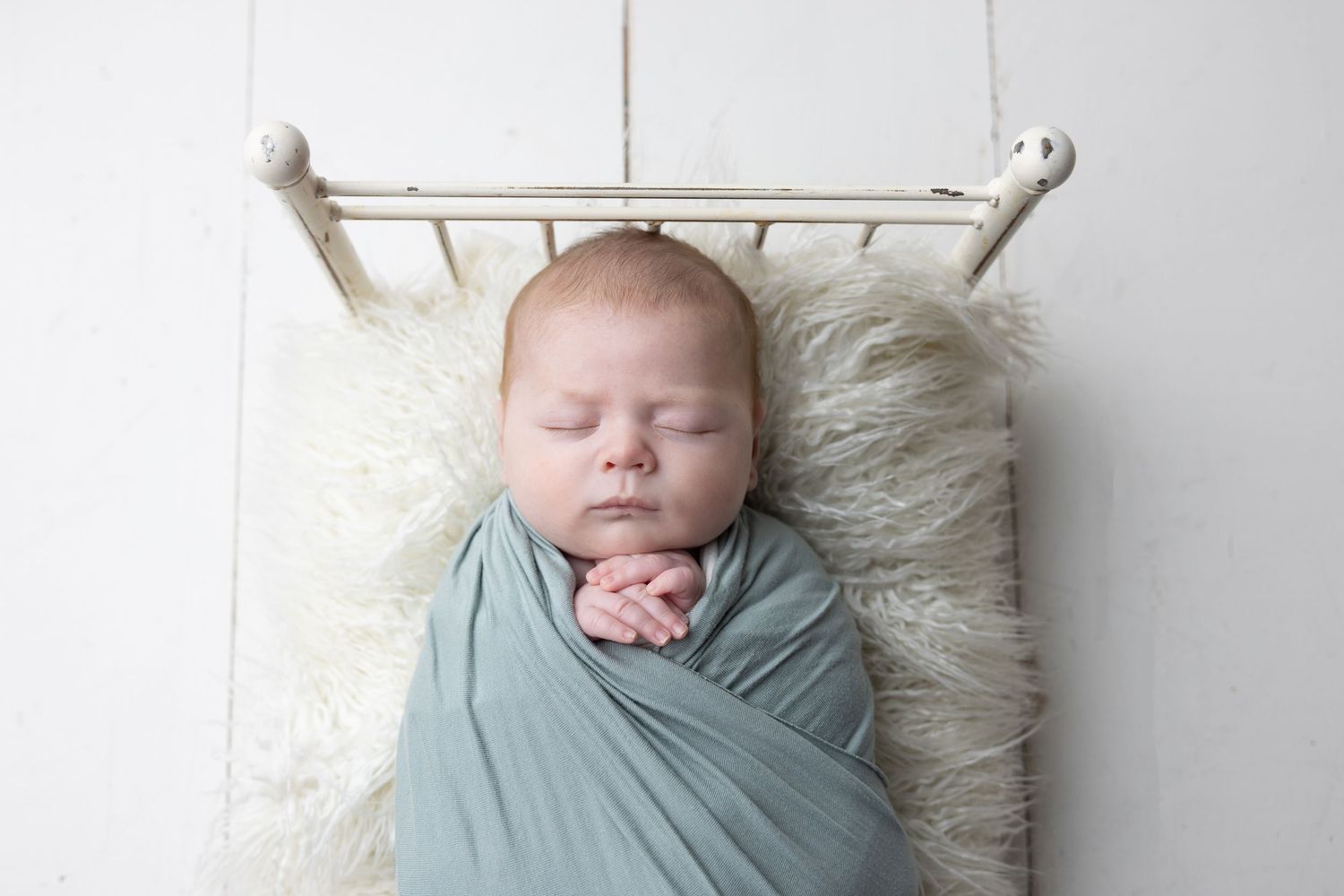 Sleeping newborn baby swaddled in a soft teal wrap, resting on a fluffy white fur blanket in a tiny white bed.