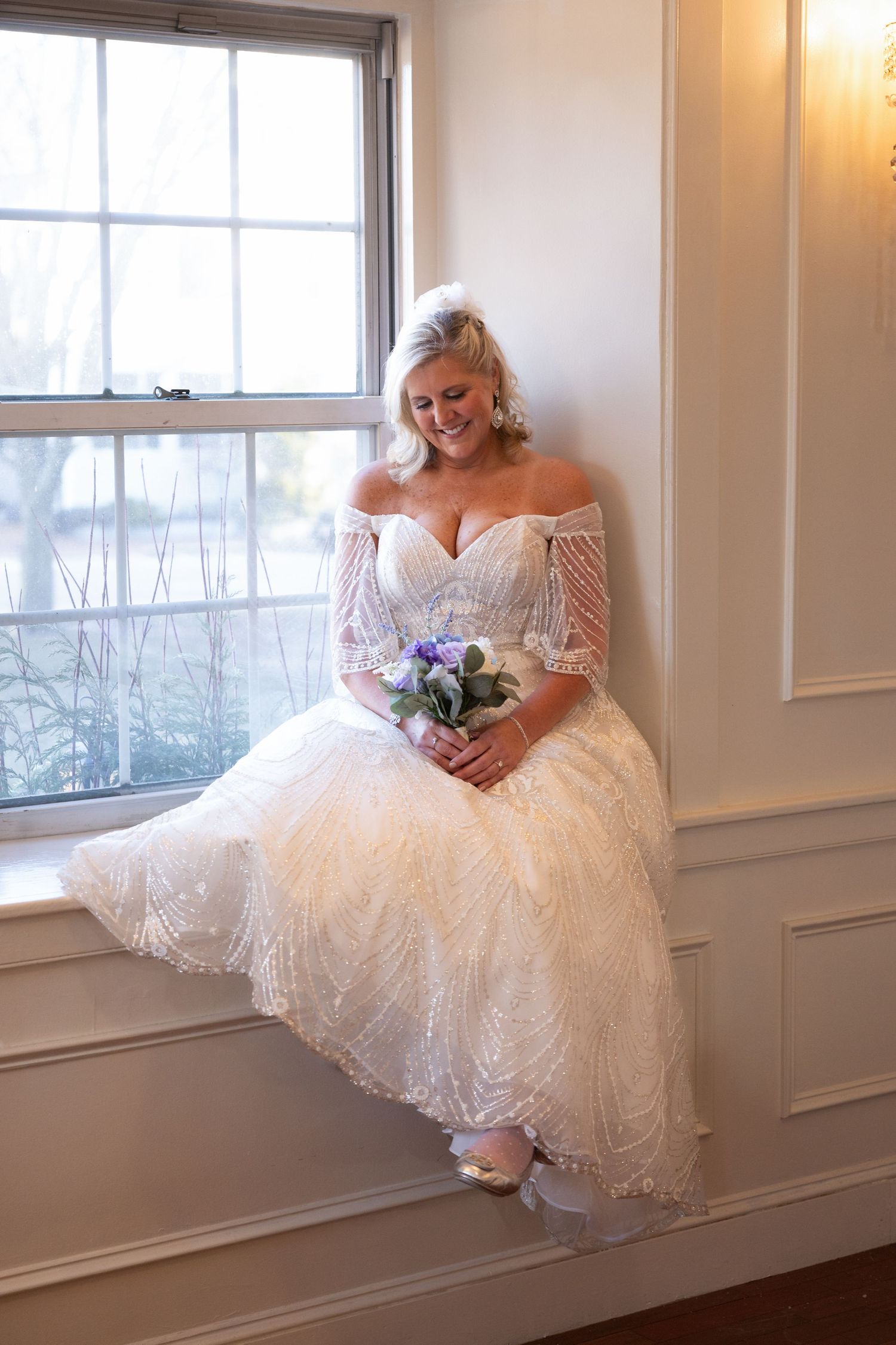 Smiling bride in off-shoulder lace gown holding purple bouquet, seated by window in elegant white room.