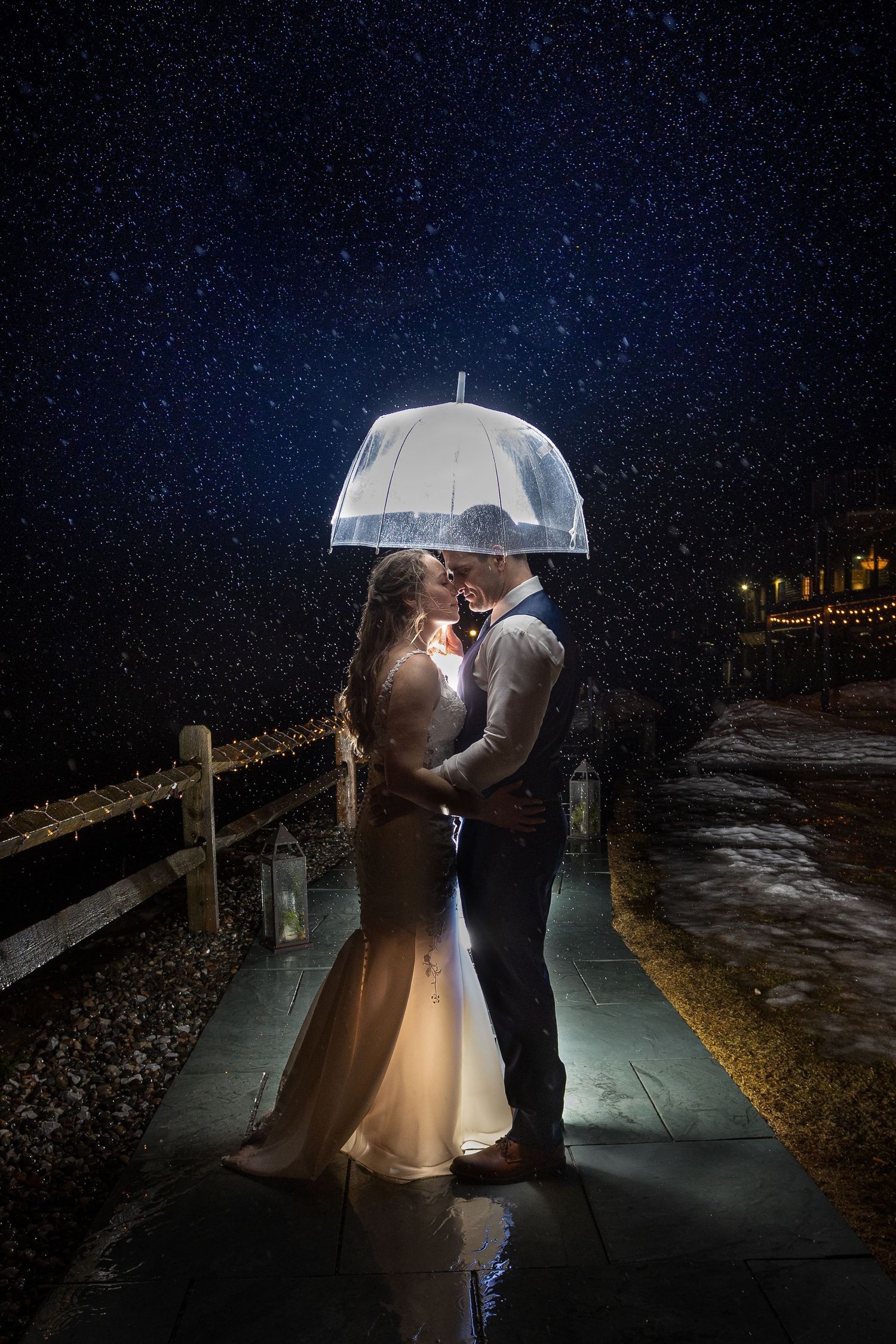 Couple sharing a romantic kiss under a glowing umbrella in the rain at night, dressed in wedding attire.