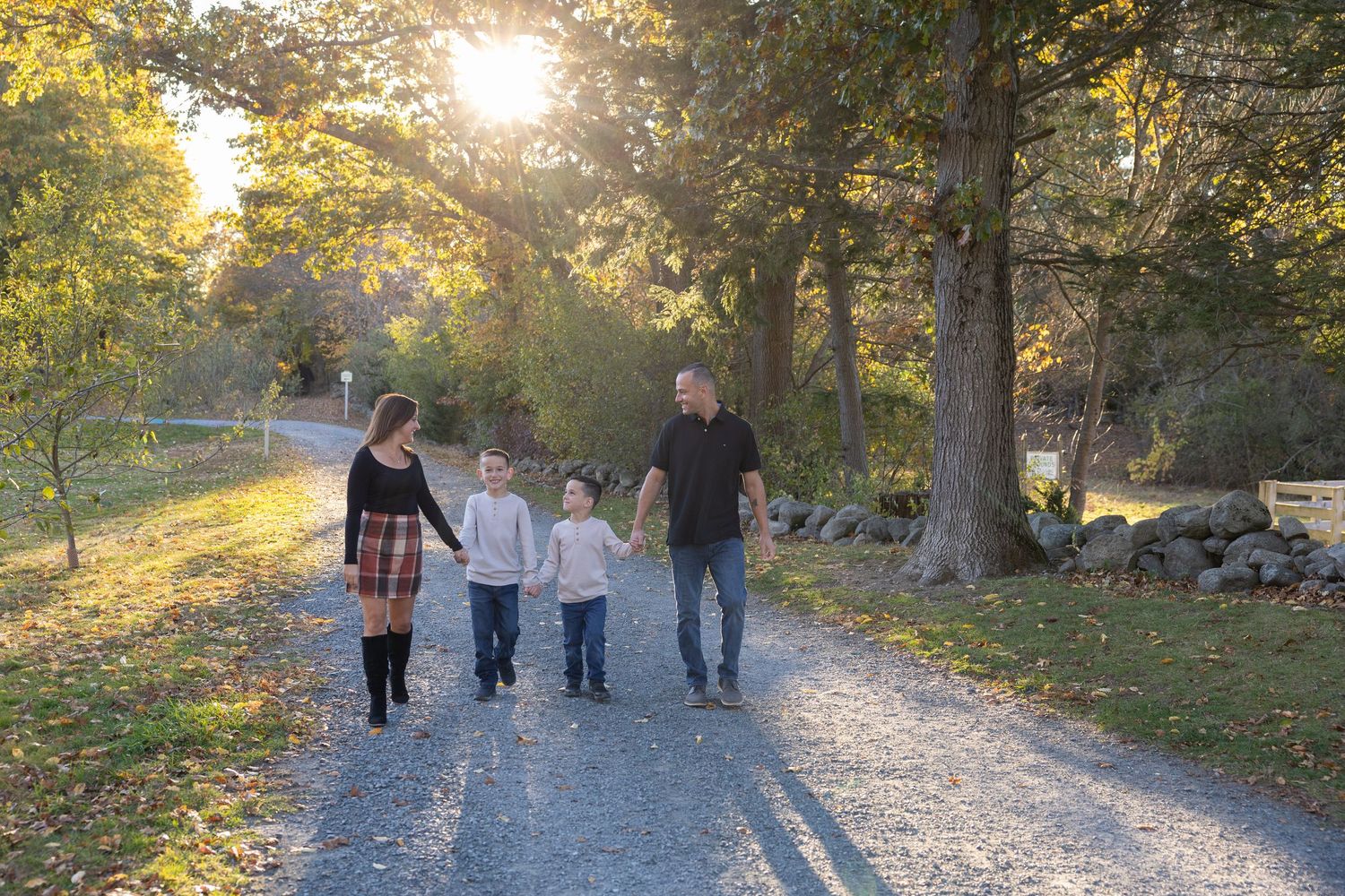 Family of four walking hand-in-hand down sunlit gravel path surrounded by colorful autumn trees.