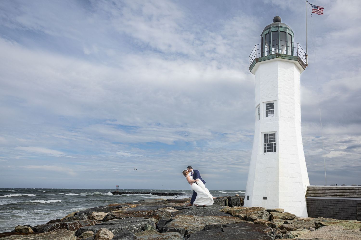 Couple embracing on rocky shore beside a white lighthouse with American flag, ocean waves in background.