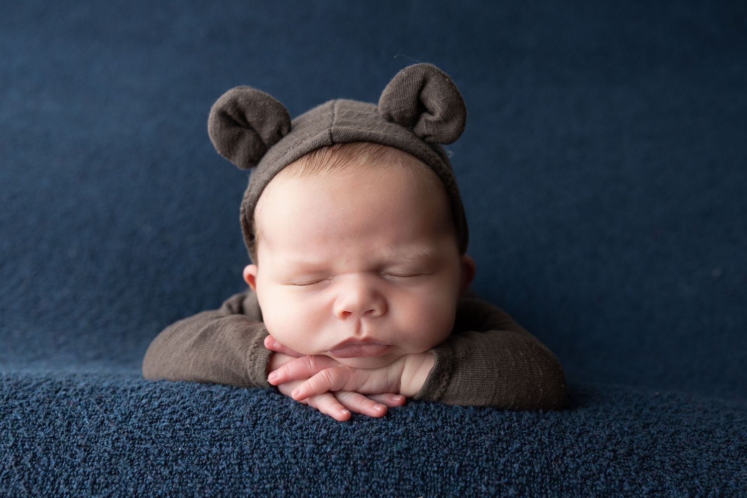 Sleeping newborn baby wearing a brown bear ear bonnet, resting chin on hands against a blue background.