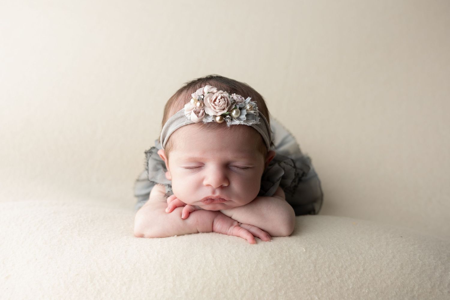 Sleeping newborn baby girl in gray outfit and floral headband posed with hands under chin on cream blanket.
