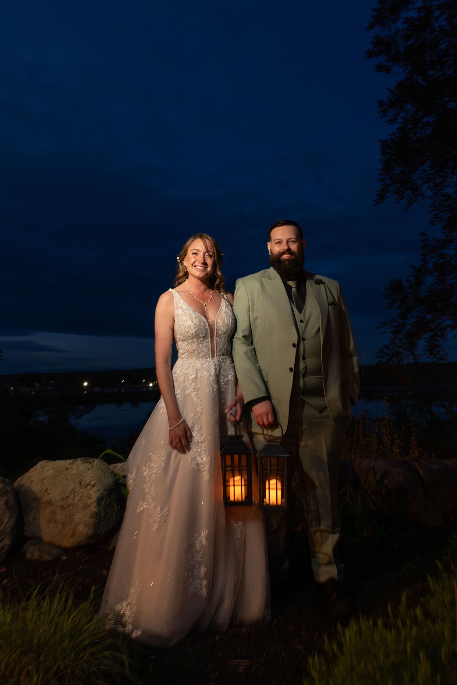 Bride and groom pose with a glowing lantern at twilight, overlooking a scenic waterway at their outdoor wedding.