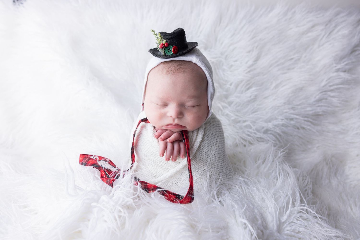 Sleeping newborn baby wrapped in white swaddle wearing tiny black top hat on fluffy white fur backdrop.