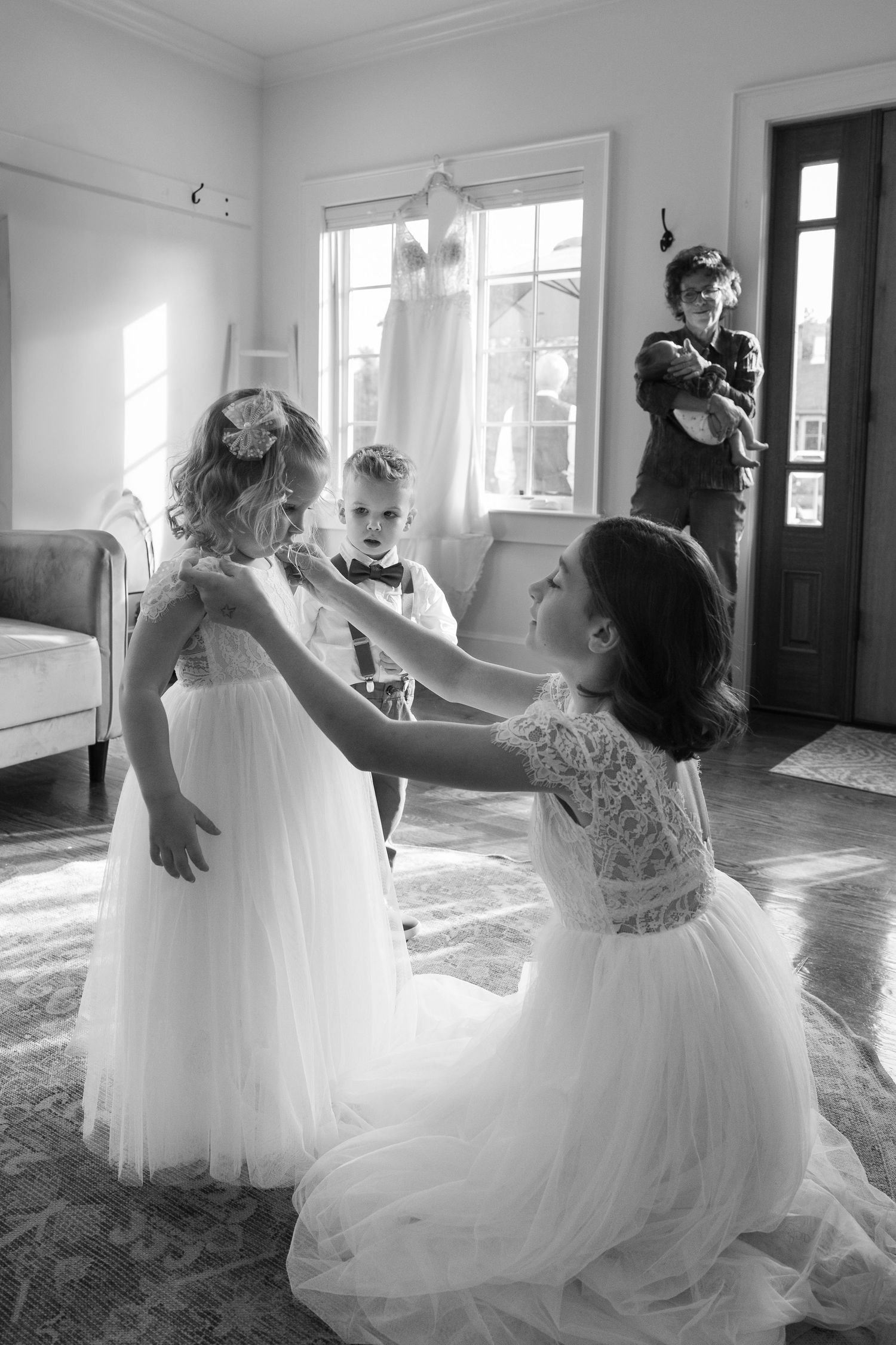 Black and white photo of a bride adjusting a flower girl's dress while children play in a sunlit room.