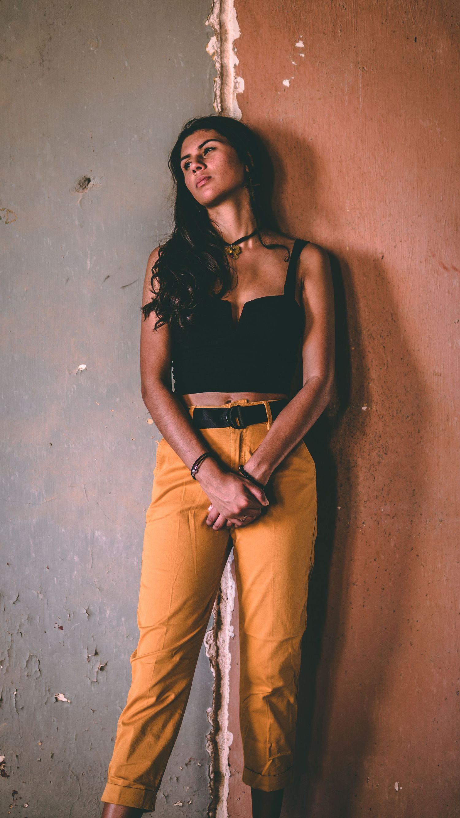 Woman in black crop top and yellow pants leaning against a weathered wall in moody lighting.