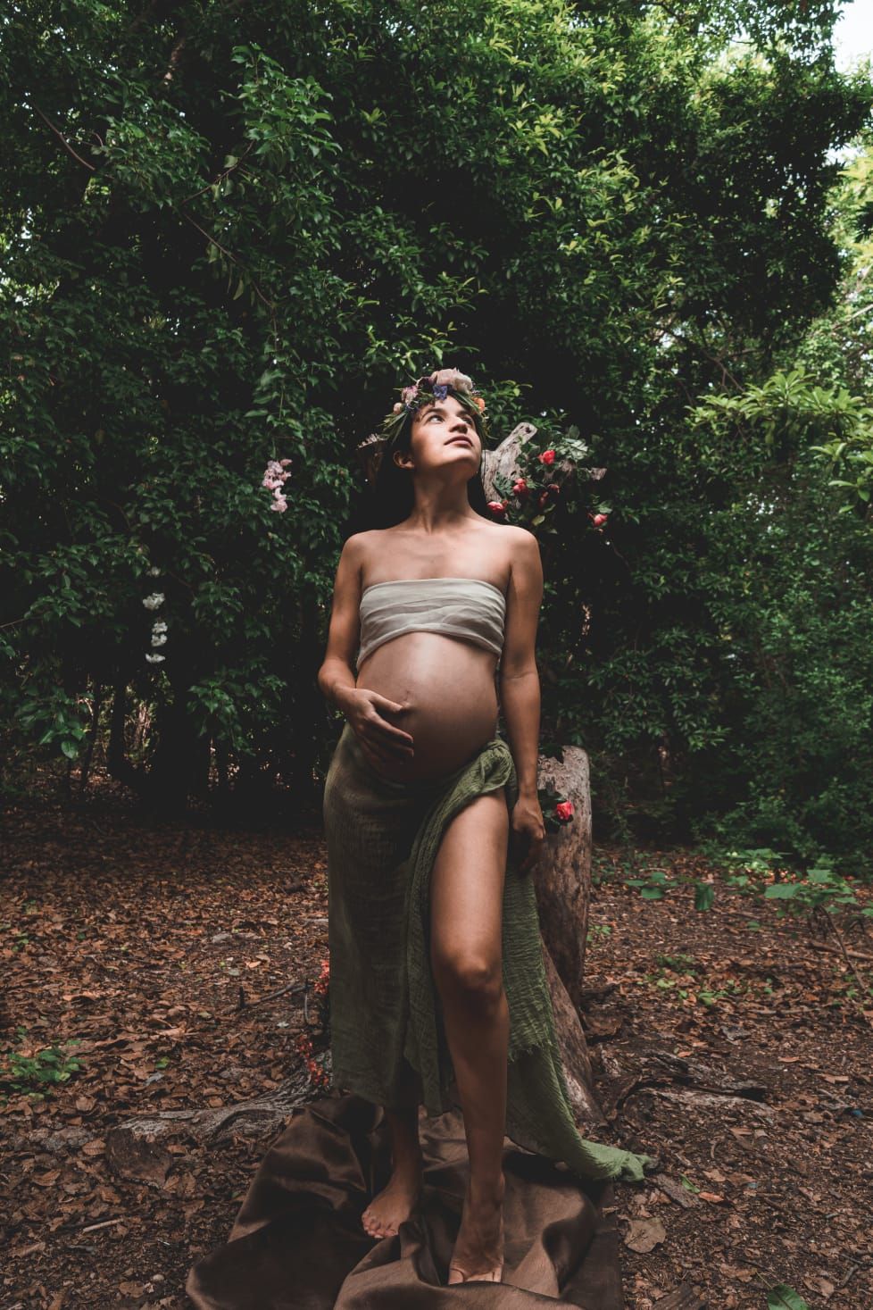 Pregnant woman in green wrap and floral crown posing on a tree stump in a lush forest setting.