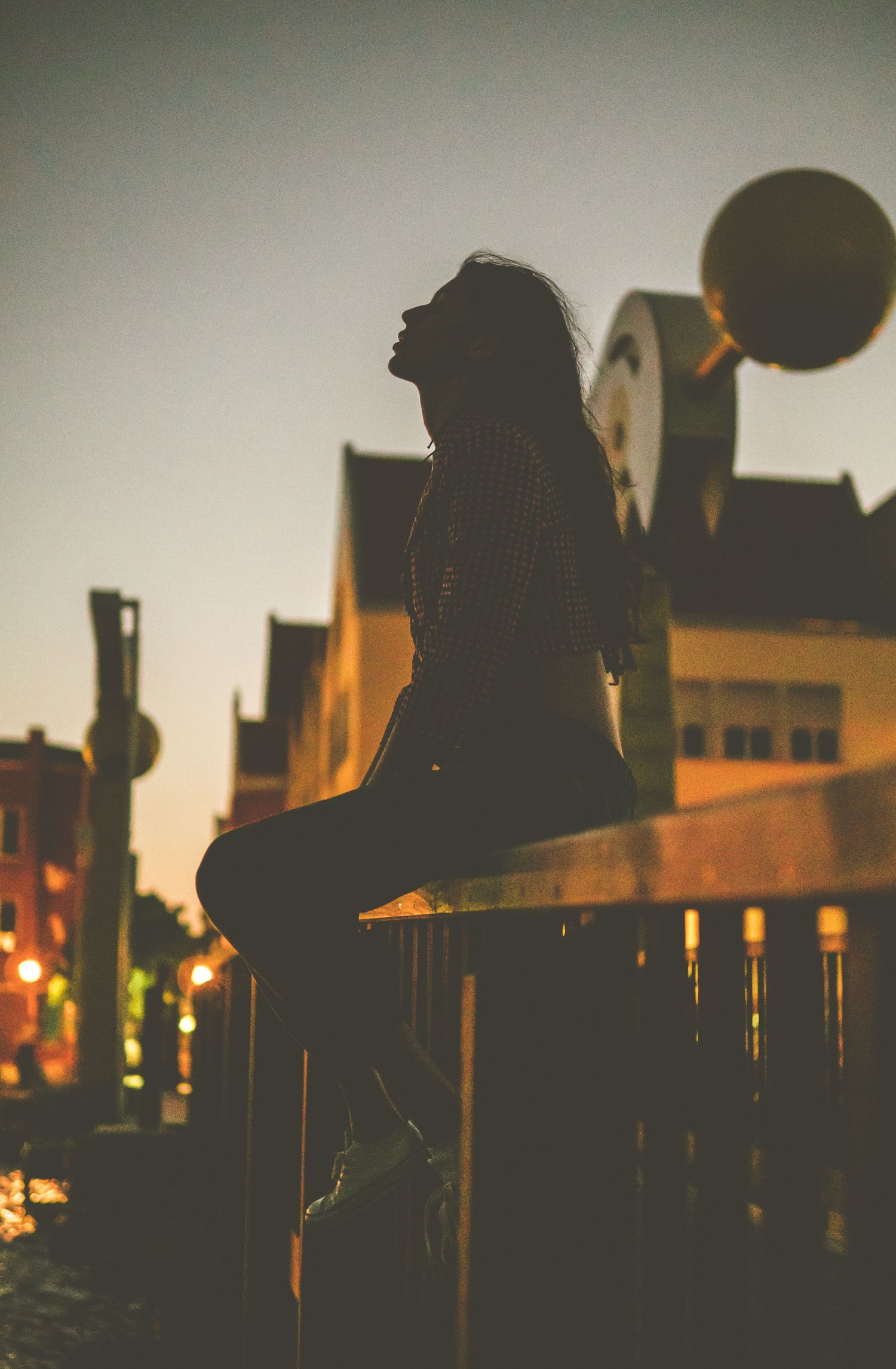Silhouette of a woman sitting on a railing at sunset in an urban city street setting.