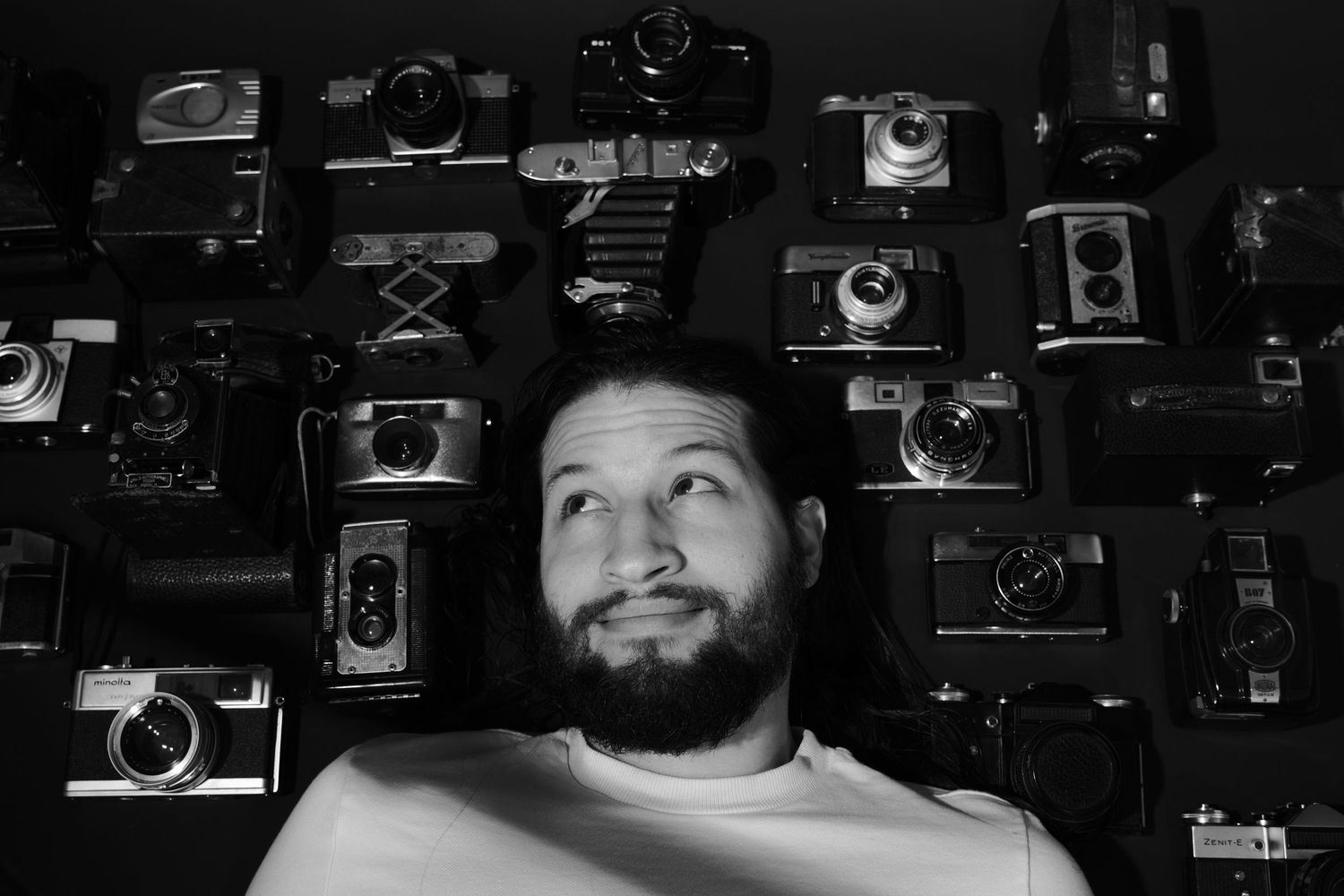 Black and white portrait of bearded man surrounded by vintage camera collection on wall.