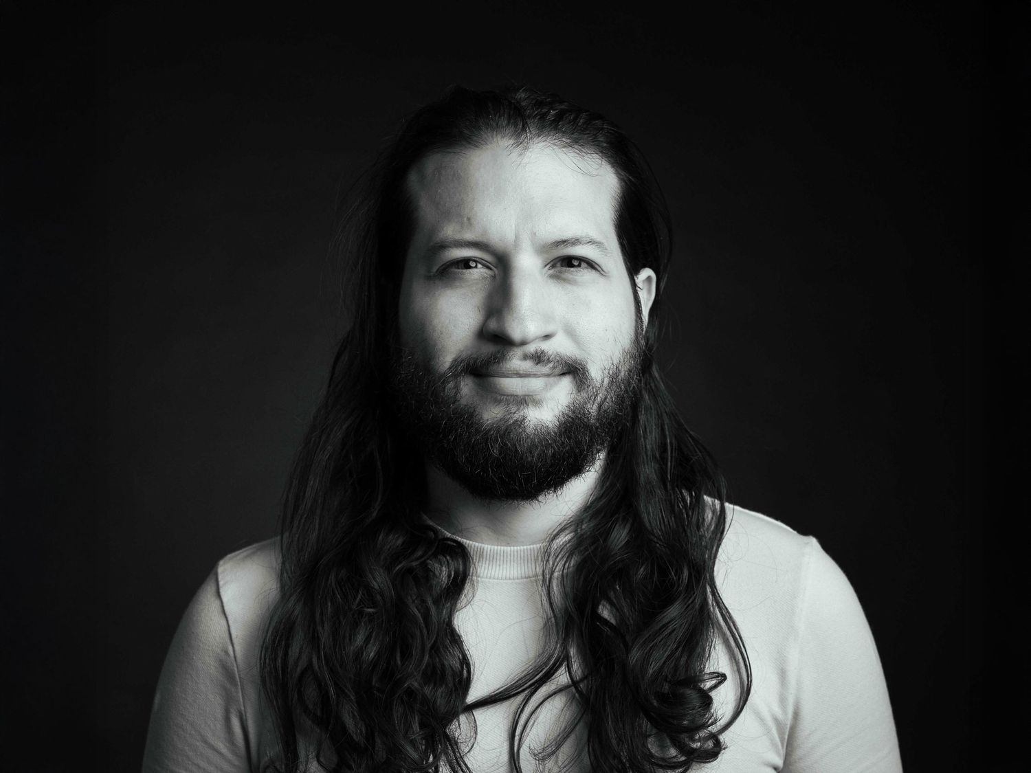 Black and white portrait of bearded man with long hair against dark background.