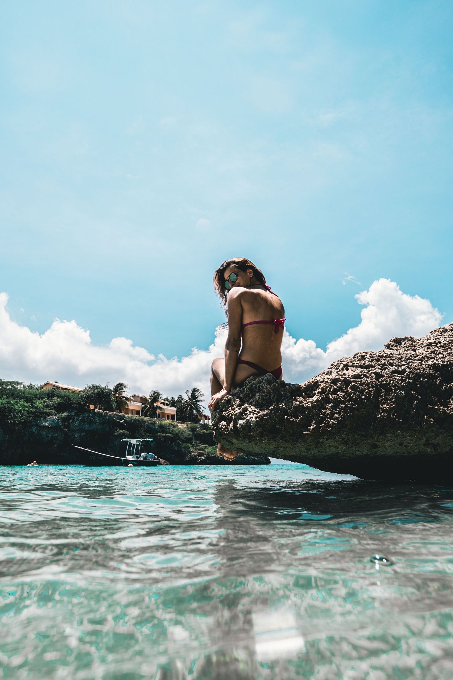Woman in red bikini sitting on coastal rocks photographed from water level under a blue sky.
