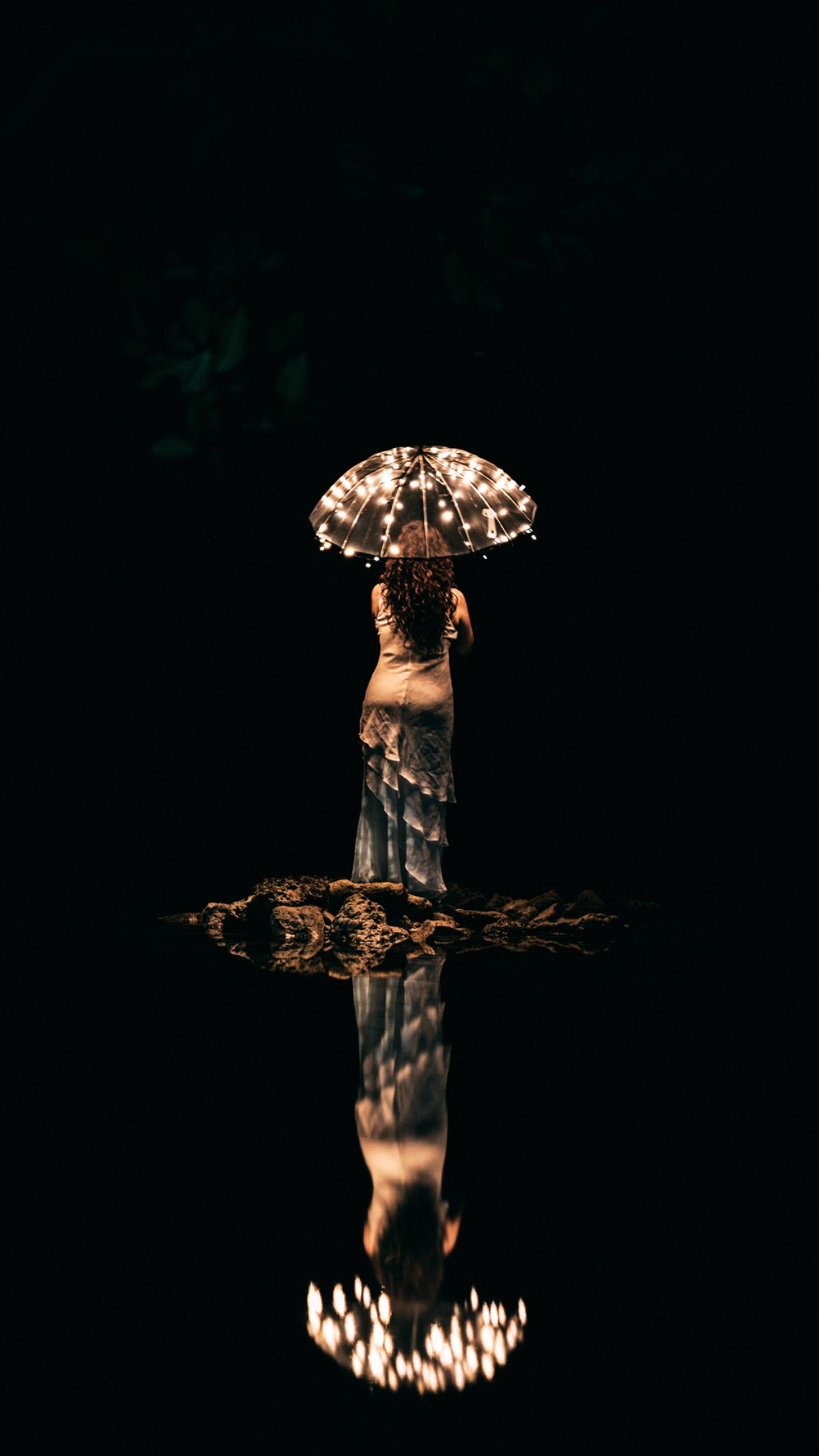 Woman in a white dress holding a lit umbrella reflected in dark water at night.