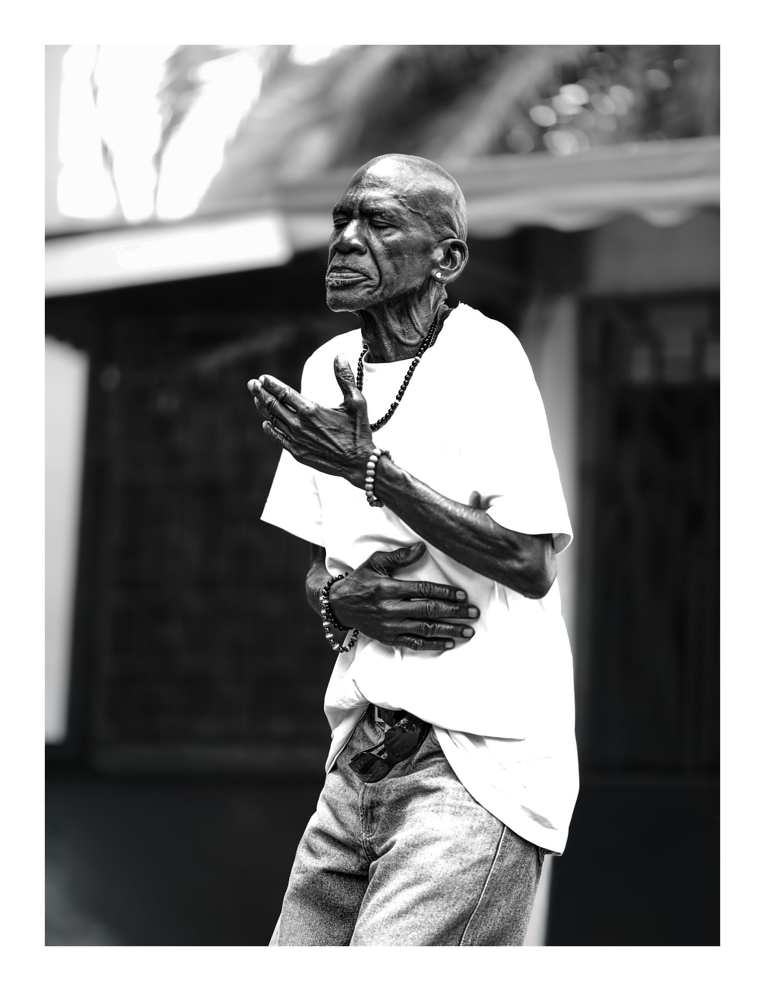 Black and white portrait of elderly man with prayer beads gesturing with hands outdoors.