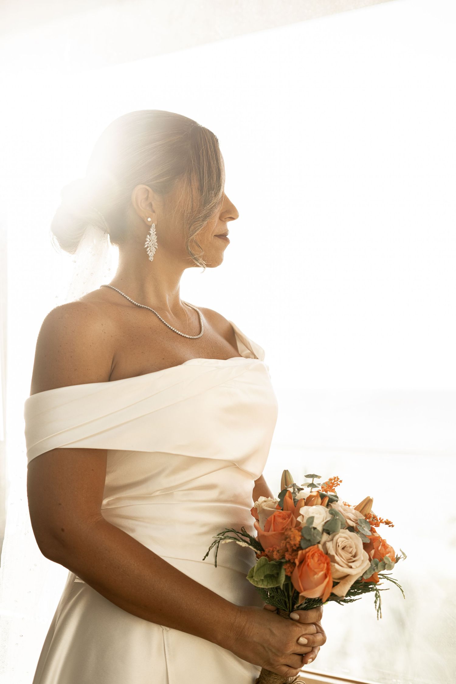 Bride in an off-shoulder white gown holding orange roses bouquet backlit by bright window light.