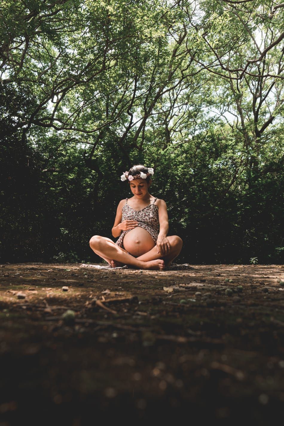 Pregnant woman with flower crown sitting cross-legged on forest ground surrounded by green trees.