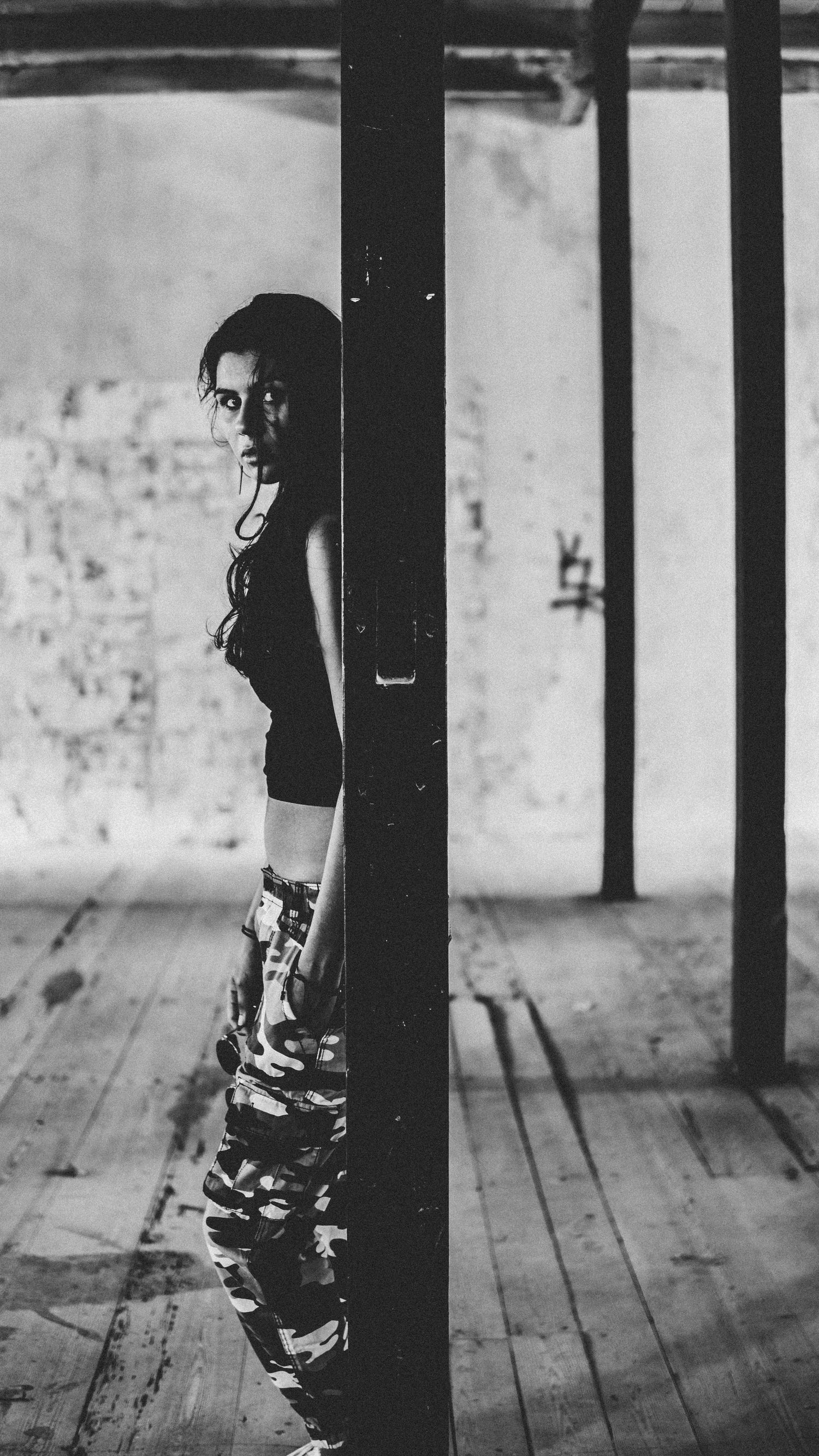 Black and white photo of a young woman in camo pants leaning against a pillar in an abandoned building.