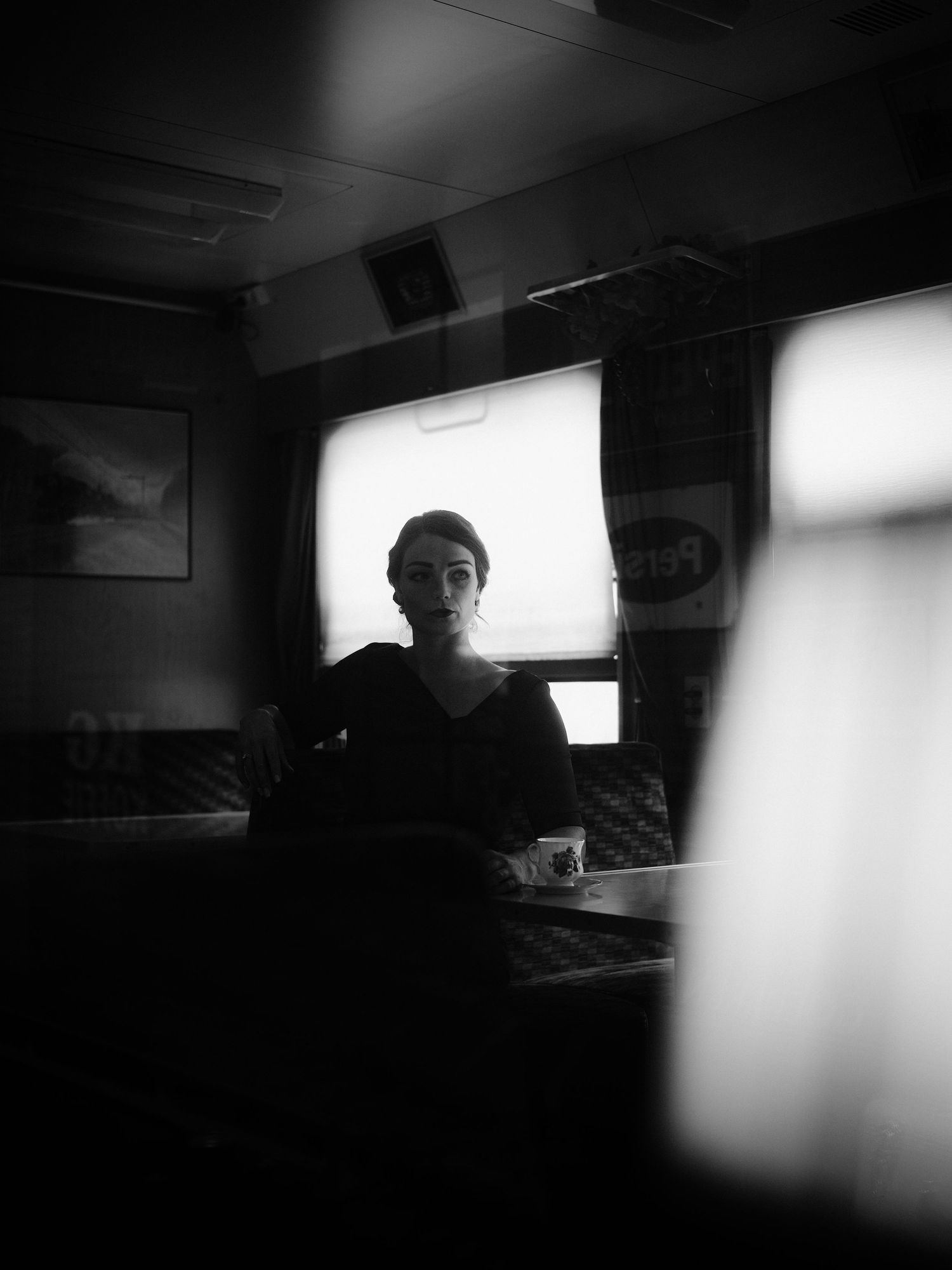 Black and white portrait of a woman sitting alone in a dimly lit diner booth near a bright window.