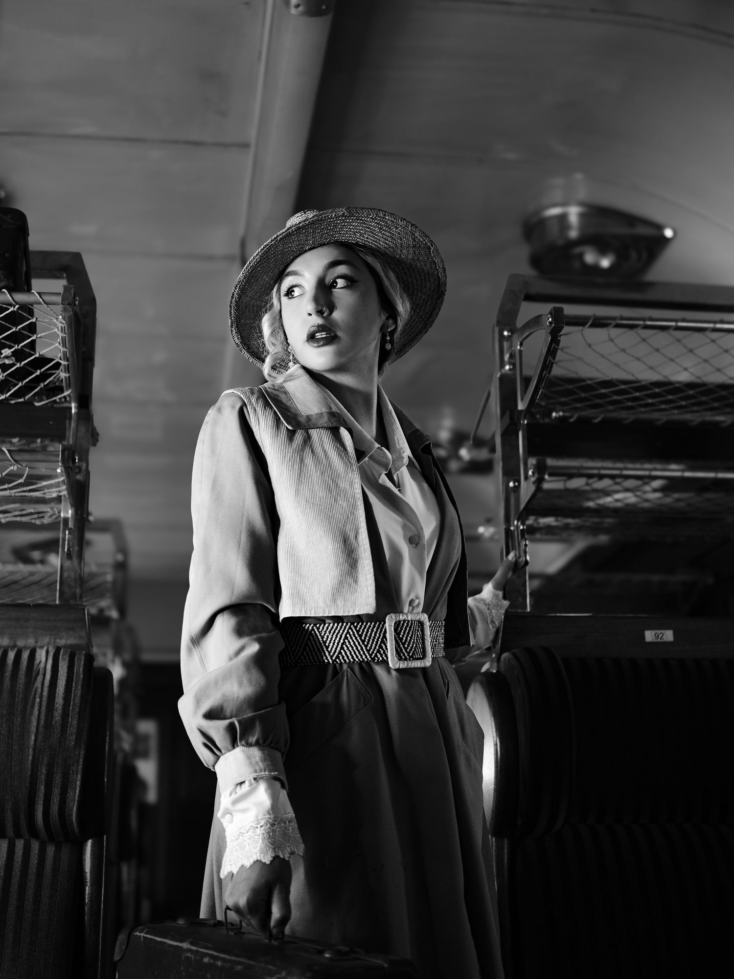 Black and white vintage-style portrait of woman in hat and trench coat with luggage.