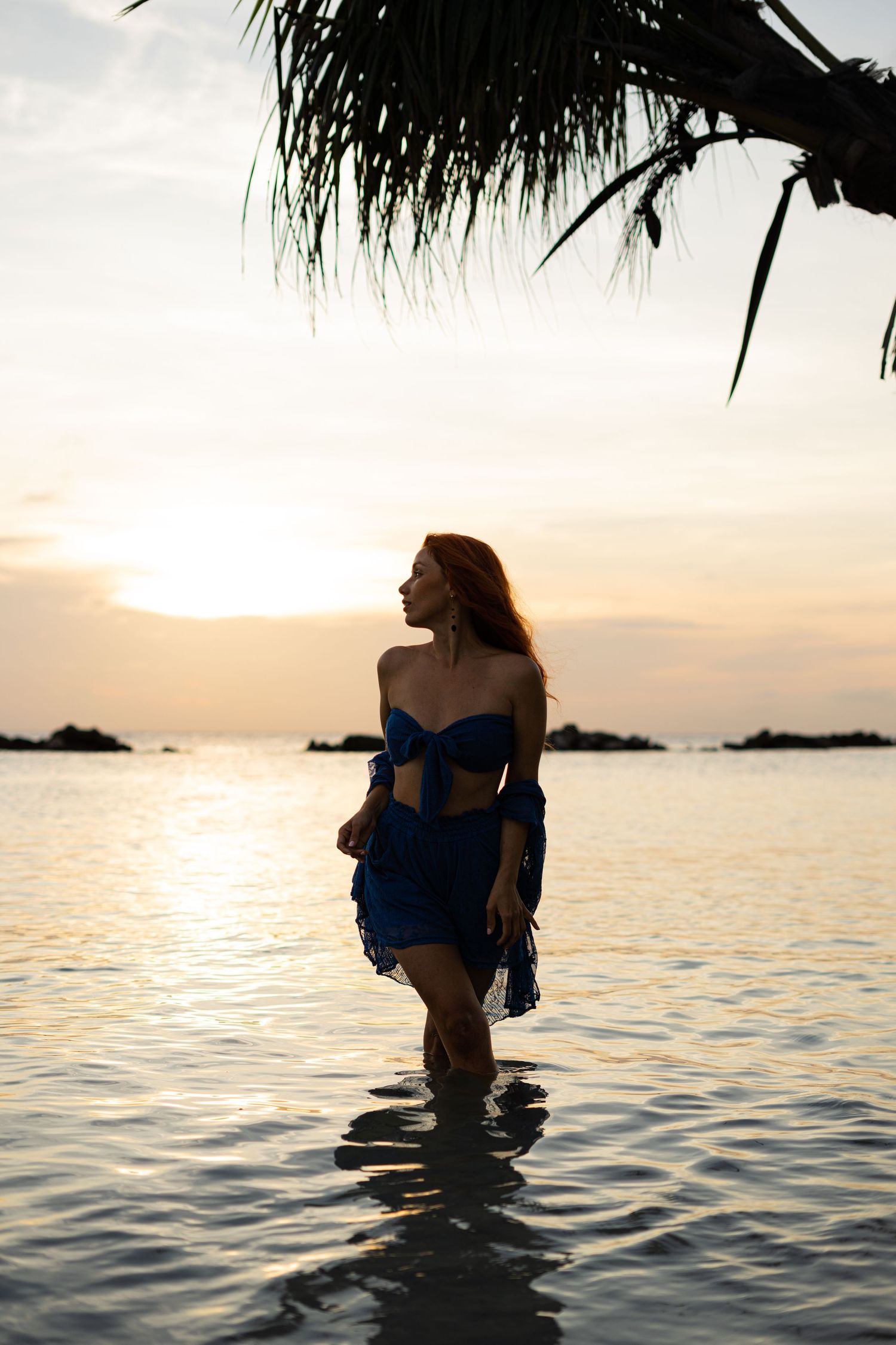 Woman in blue two-piece outfit wades through shallow ocean water during a vibrant tropical sunset.