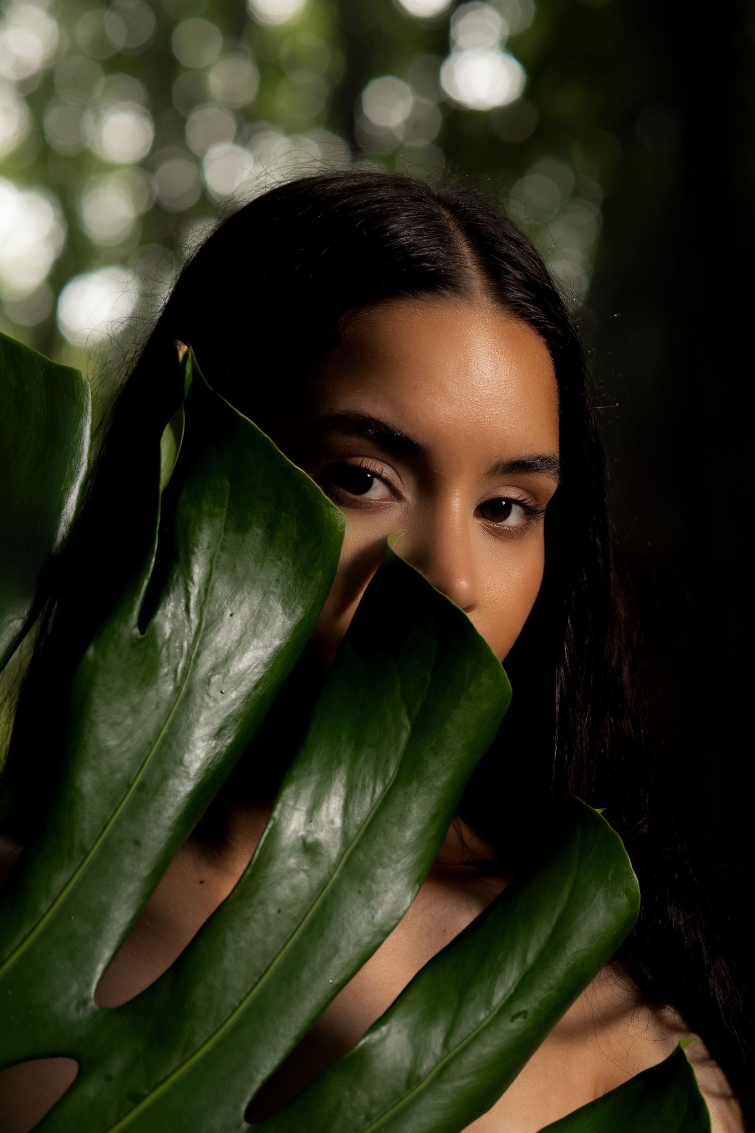 Close-up portrait of a young woman partially hiding her face behind a large tropical green leaf.