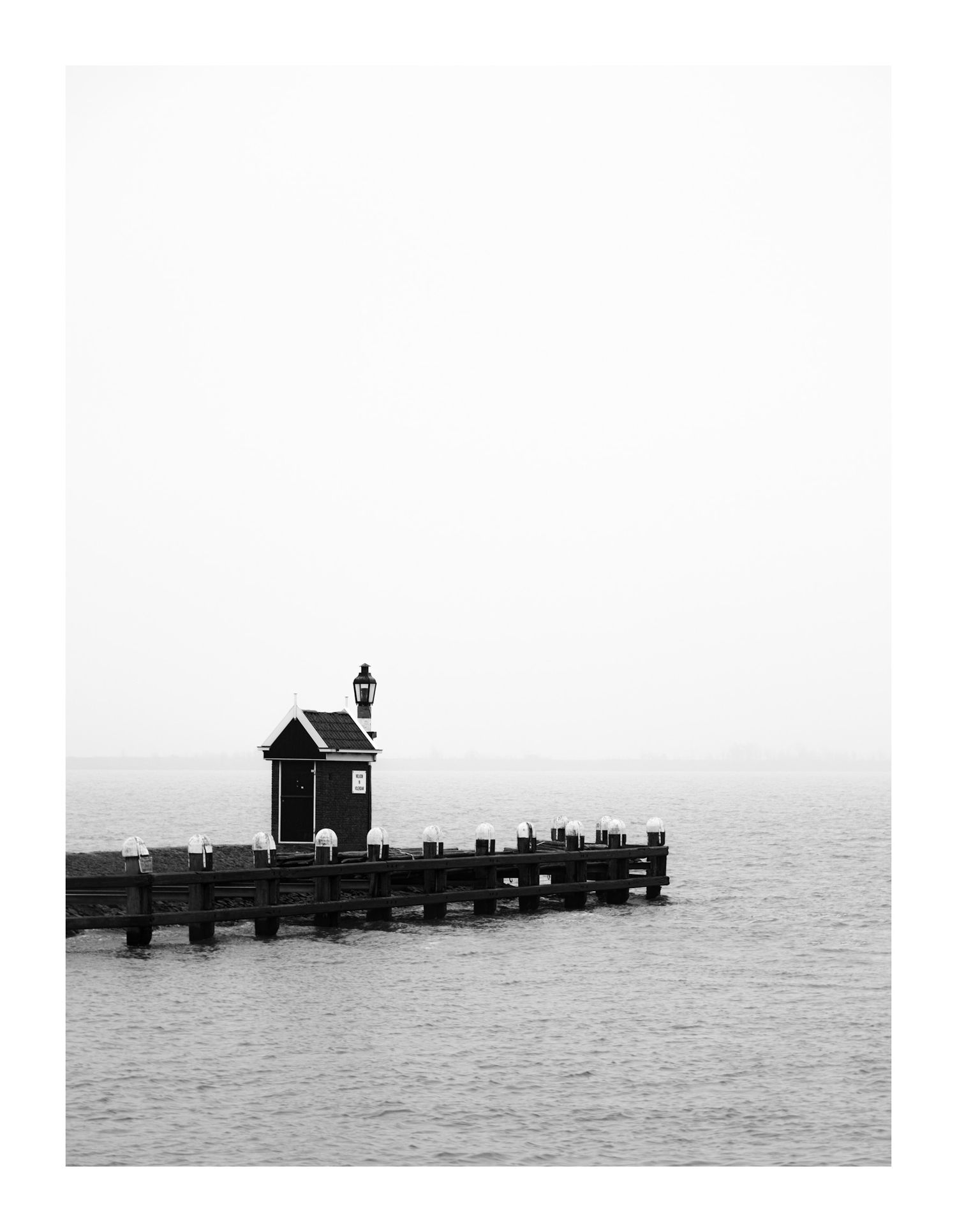 Lonely wooden pier with small hut and lamp extending into calm misty sea, black and white.