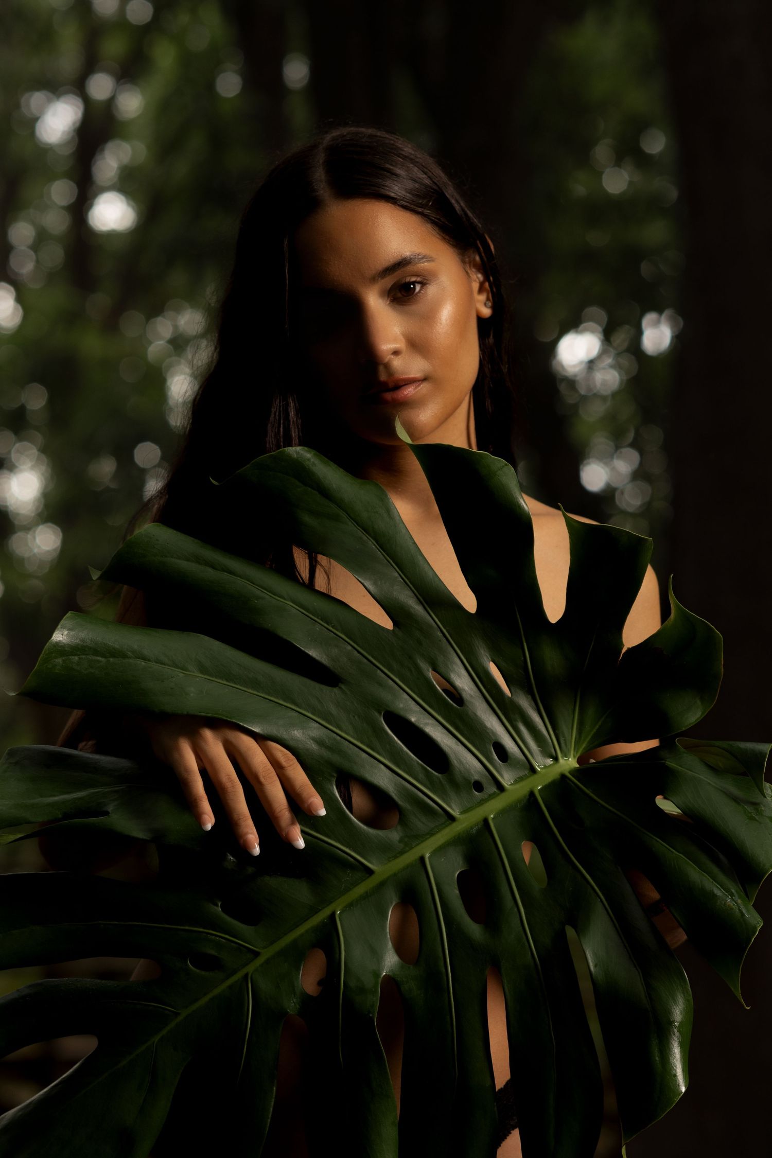 Brunette woman holds large monstera leaf in dark forest setting for nature portrait photography.