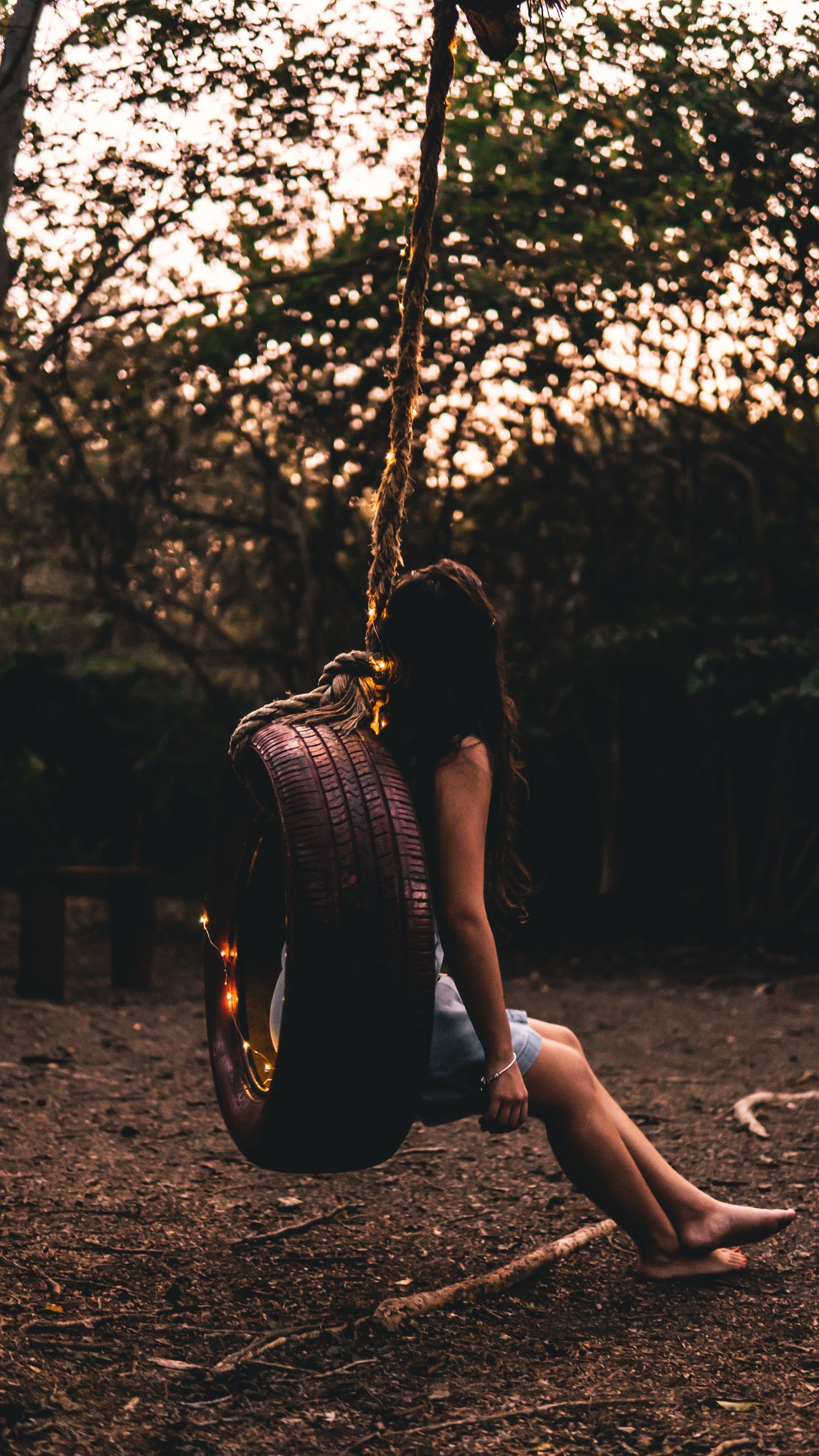 Young woman sitting on a tire swing at golden hour surrounded by glowing bokeh tree lights.