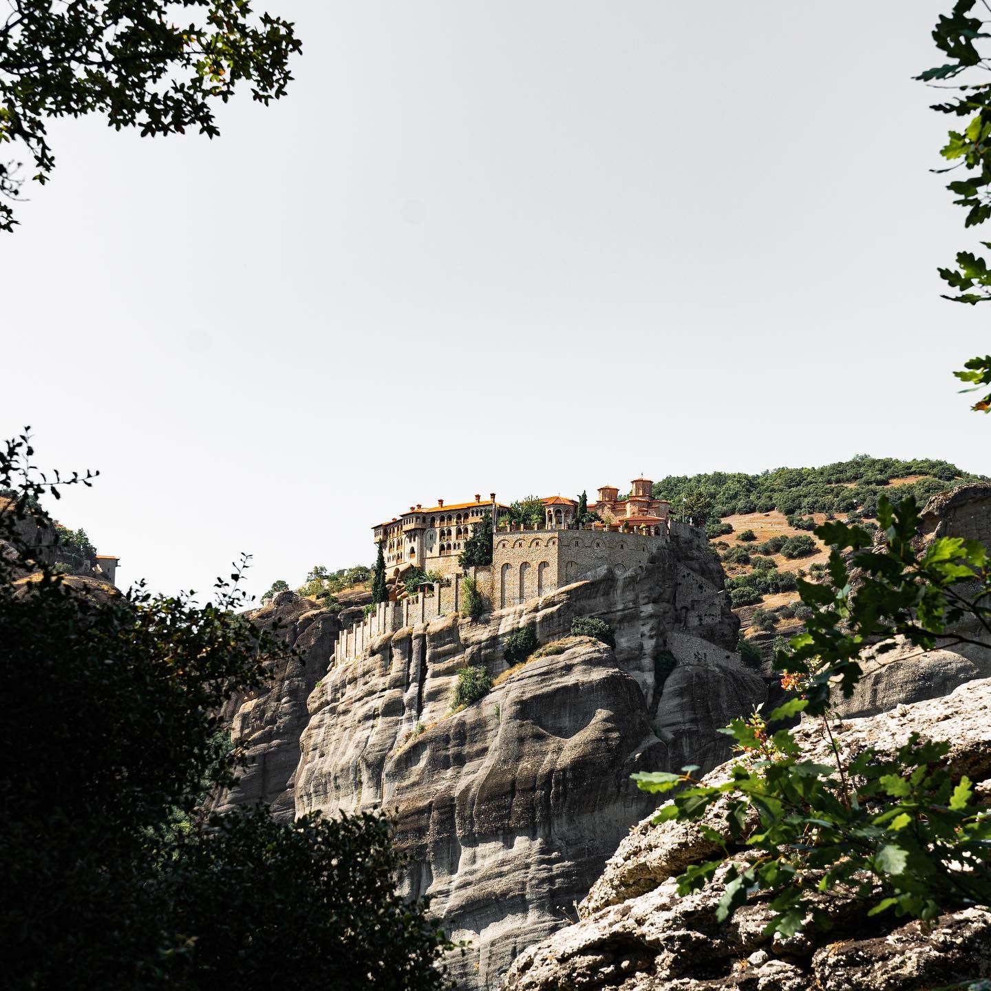 Ancient monastery perched atop dramatic rocky cliffs at Meteora, Greece, surrounded by lush green trees under a hazy sky.