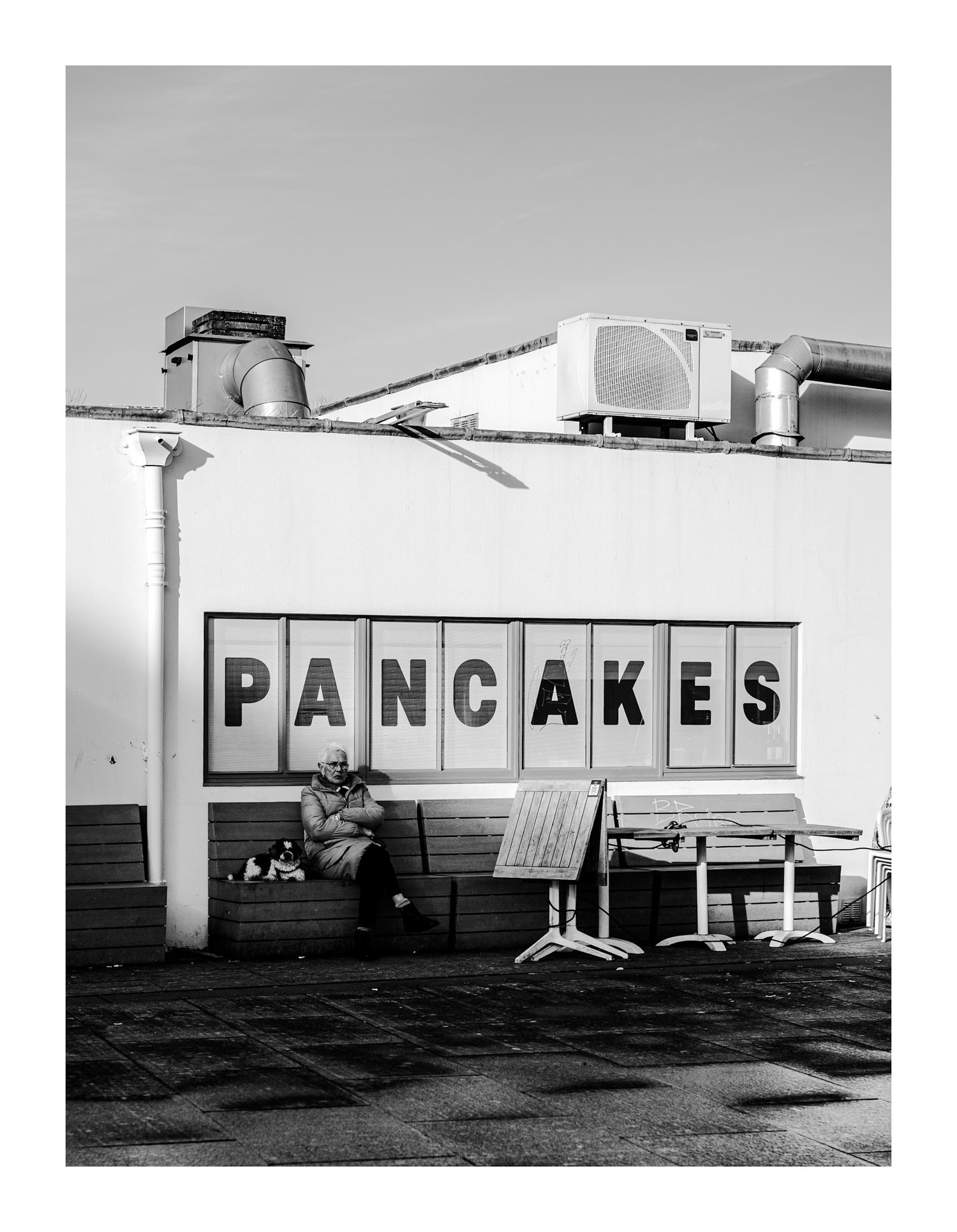 Person sitting alone beneath large Pancakes sign on building exterior, black and white photo.