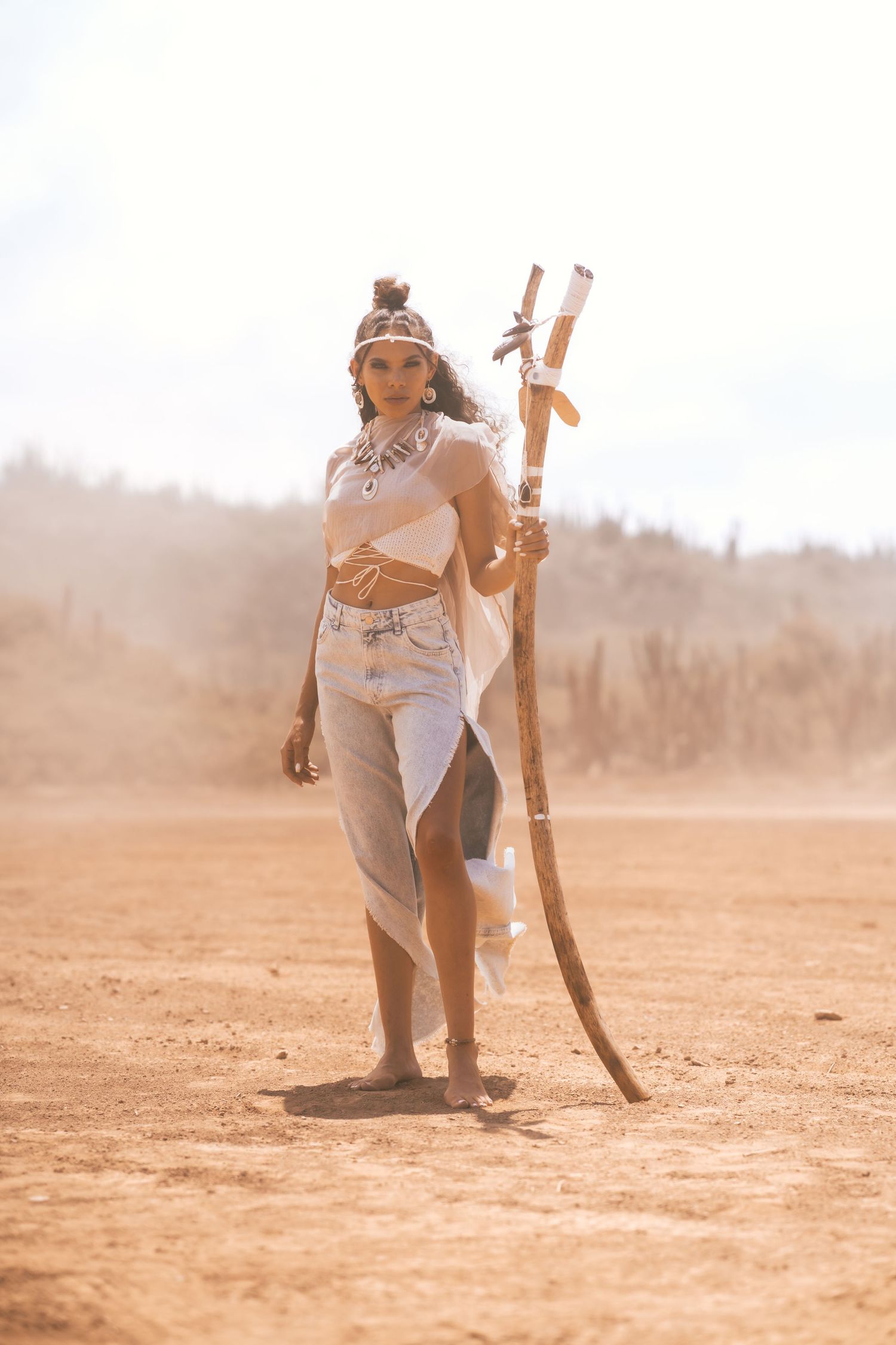 Woman in tribal boho outfit holding wooden staff in sandy desert landscape.